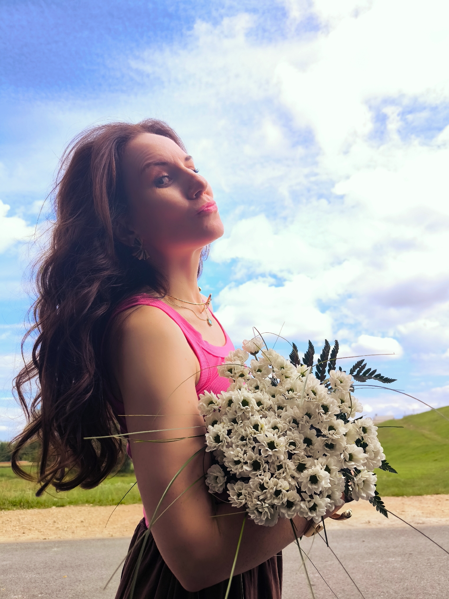 A woman with long, dark, wavy hair stands outdoors, holding a bouquet of white daisies. She wears a bright pink tank top and a dark skirt. Her head is tilted back, and she looks upwards towards the sky. The background features a bright blue sky with fluffy white clouds, a green grassy hill, and a dirt road. The image appears to have been taken on a sunny day. The woman is positioned in the foreground, with the natural landscape forming the backdrop. There is no visible indication of the city of Riga, Latvia in the image.