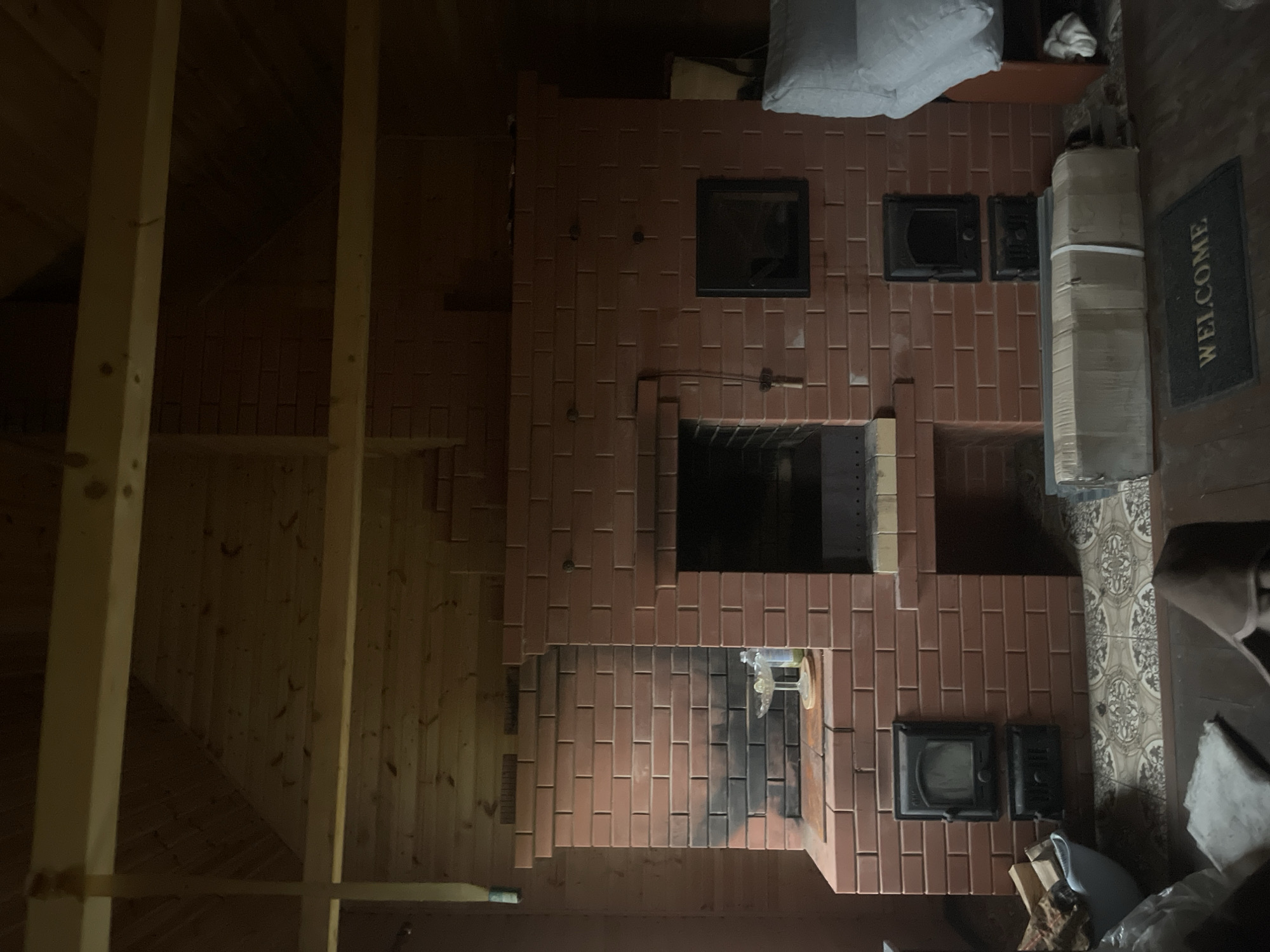 This media file displays an interior scene of a room, likely a house, in Krasnoe, Russia. The focal point of the image is a large, unfinished brick stove or fireplace that occupies a significant portion of the frame. The stove is constructed from reddish-brown bricks and features several openings, including a main firebox and what appear to be smaller access doors or vents. Some parts of the stove show signs of soot or charring, suggesting recent use or ongoing construction.
To the left of the stove, the wall is made of light-colored wooden planks, giving a rustic feel to the space. Exposed wooden beams are also visible, suggesting that this might be an attic or a room with an exposed ceiling structure. To the right of the stove, the wall features patterned tiles and a dark welcome mat with the word "WELCOME" printed on it in white. Various objects are scattered around, including stacks of boxes, a white pillow or bag, and some firewood. The lighting in the room is dim, creating a somewhat shadowed atmosphere, and it is difficult to determine the time of day or weather conditions outside. No people are visible in the image, and the overall impression is one of a work-in-progress or a utilitarian space.