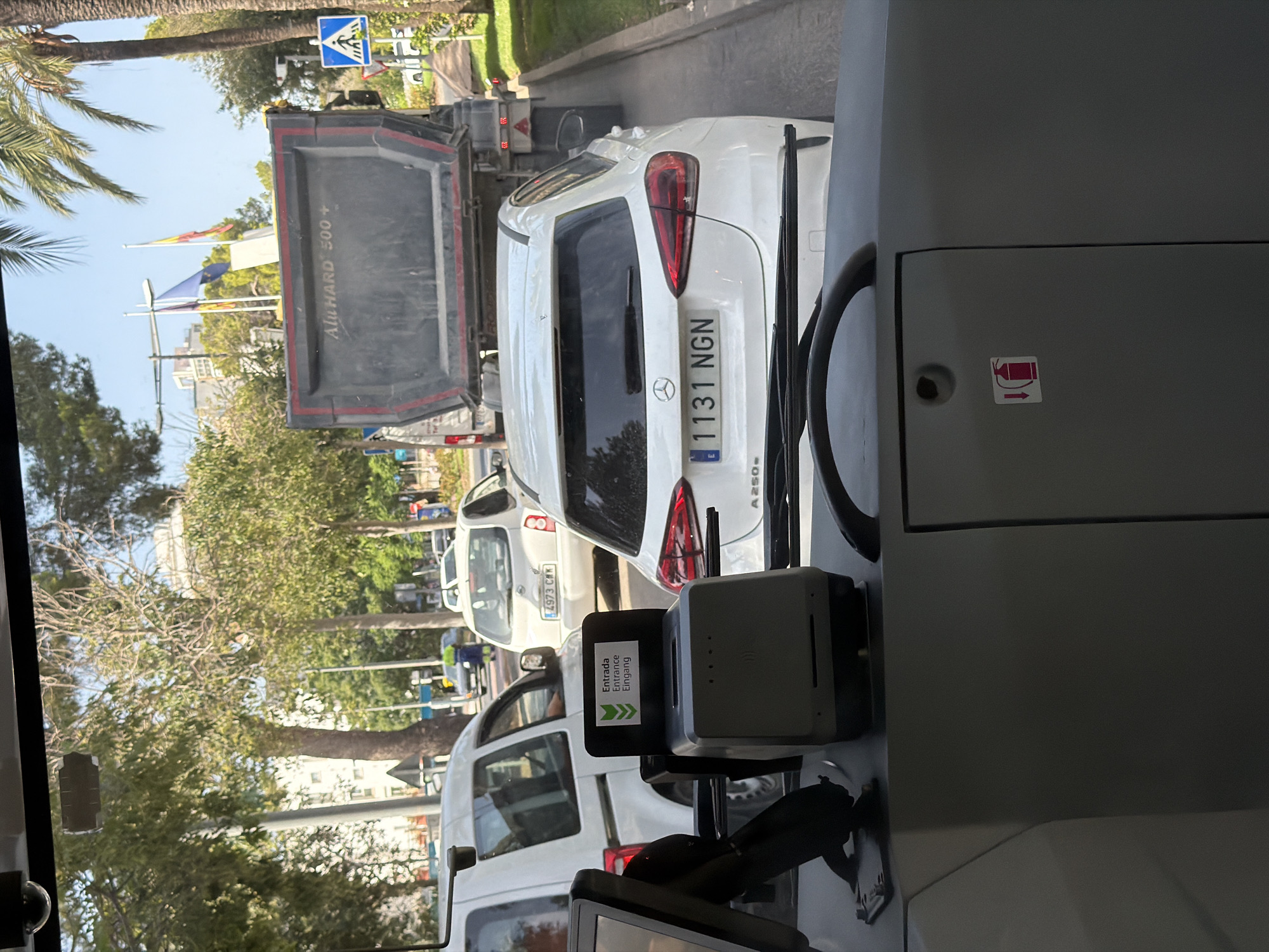 The image shows a view from inside a vehicle, looking out through the windshield. In front of the vehicle, there is a ticket machine with an "Entrada Entrance Eingang" sign. Beyond the ticket machine, traffic is visible. A white Mercedes-Benz A-Class car with license plate "1131 NGN" is directly in front. To the left of the Mercedes is a white panel van. Further ahead, a large black dump truck with "AluHARD 500+" written on it is stopped. Other vehicles, trees, buildings, and a pedestrian crossing sign are also visible in the background. The scene appears to be a street in Calvià, Spain, likely on a sunny day.