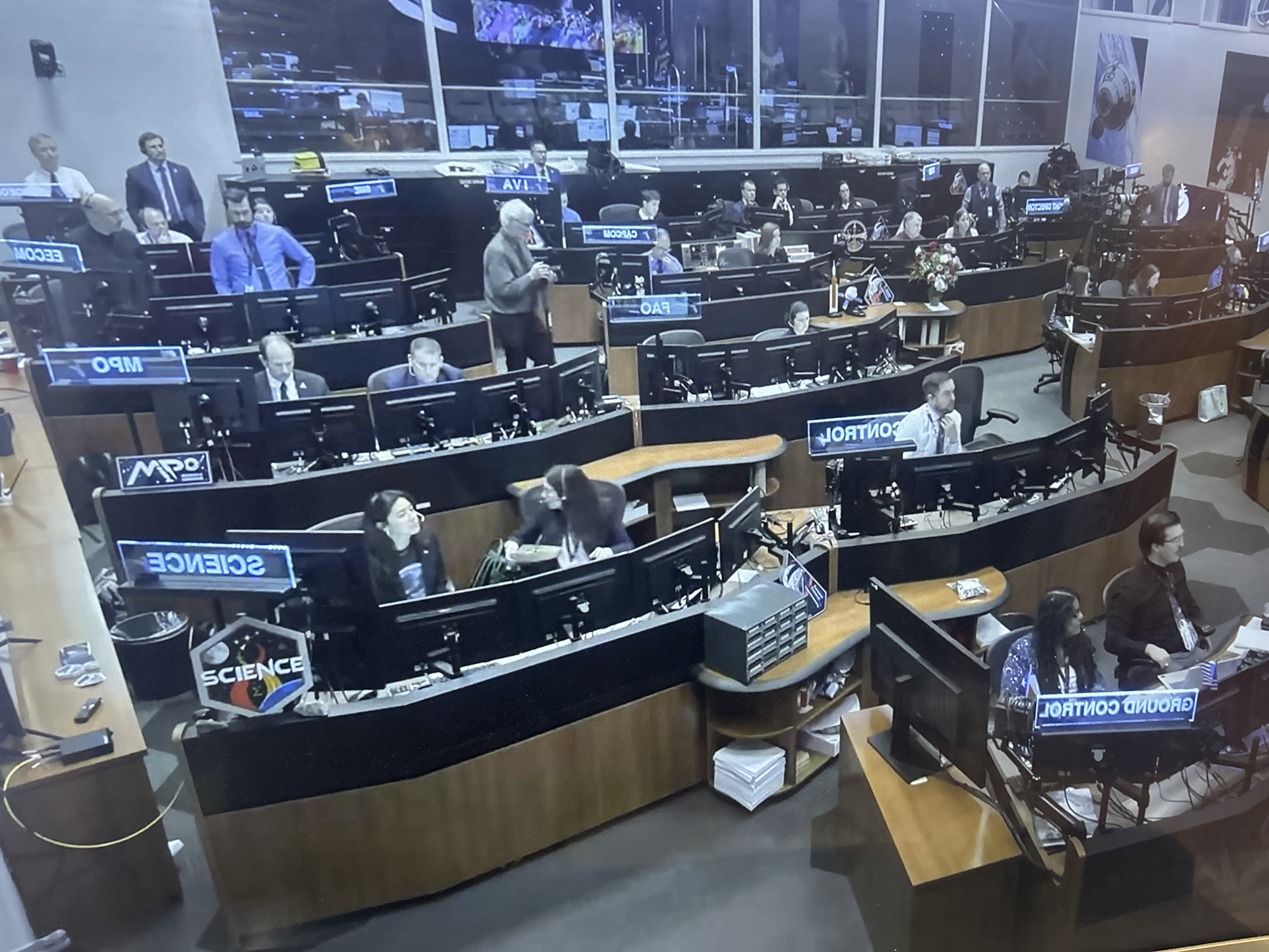 The image shows a control room filled with people working at computer stations. The stations are arranged in tiers, with labels such as "EECOM," "MPO," "AVI," "OAR," "SCIENCE," and "GROUND CONTROL" displayed on them. Many people are seated at the computers, while others are standing and observing. The environment suggests a high-tech, mission-critical operation, possibly a space mission control center, though the location is Unknown, Unknown.