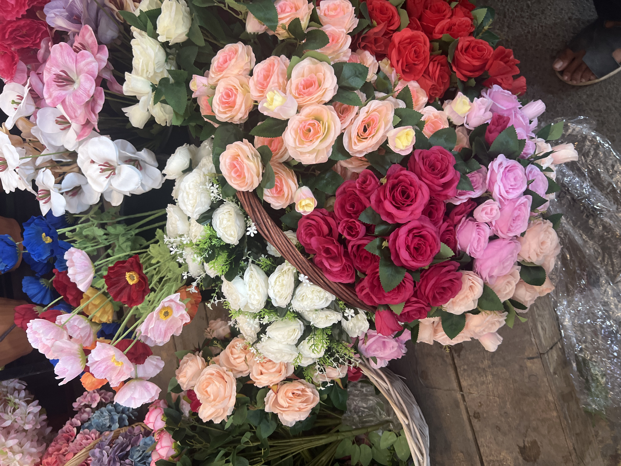 The image depicts a close-up, overhead view of a flower stall, likely in Bishkek, Kyrgyzstan, exhibiting a vibrant and diverse collection of artificial flowers. The primary focus is on a large assortment of roses in varying shades of pink, from light blush to deep fuchsia, and bold red. These roses are arranged in bouquets, some spilling out of a woven wicker basket and others placed on a wooden surface, possibly a market stall.
Interspersed with the roses are other types of artificial flowers, including white and pale pink blossoms with visible stamens, clusters of small white flowers, and larger, open-faced pink and white flowers that resemble poppies. In the background, there are hints of more flowers in shades of blue, purple, and other bright colors.
The ground surface is a mixture of weathered wooden planks, with some areas appearing to be covered by plastic sheeting that reflects light. A portion of a person's foot wearing a dark sandal is visible in the lower right corner of the frame, suggesting the presence of a shopper or vendor. The overall impression is one of abundance and color, characteristic of a bustling market.