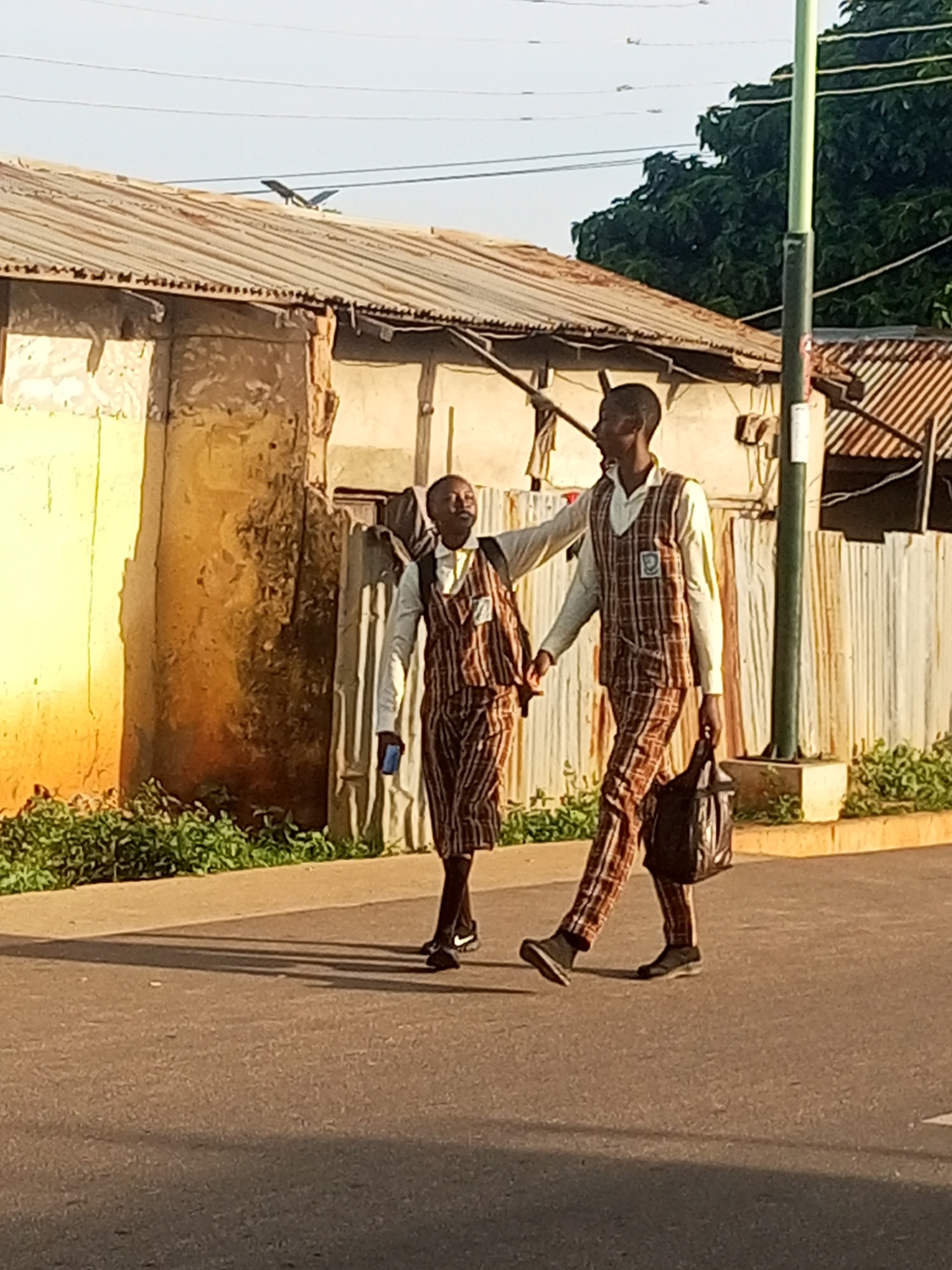 In Tumfure, Nigeria, two young males in matching plaid school uniforms walk along a paved road. The uniforms consist of a light-colored collared shirt worn under a plaid vest and plaid trousers. The male on the right is carrying a black bag in his right hand and appears to be holding the left hand of the male on the left. The male on the left is holding a blue object in his left hand. They are walking away from the viewer and towards a building with a corrugated metal roof and an orange-yellow wall. Green foliage lines the edge of the road in front of the building. A green utility pole is visible to the right of the males. The sky is clear and light.