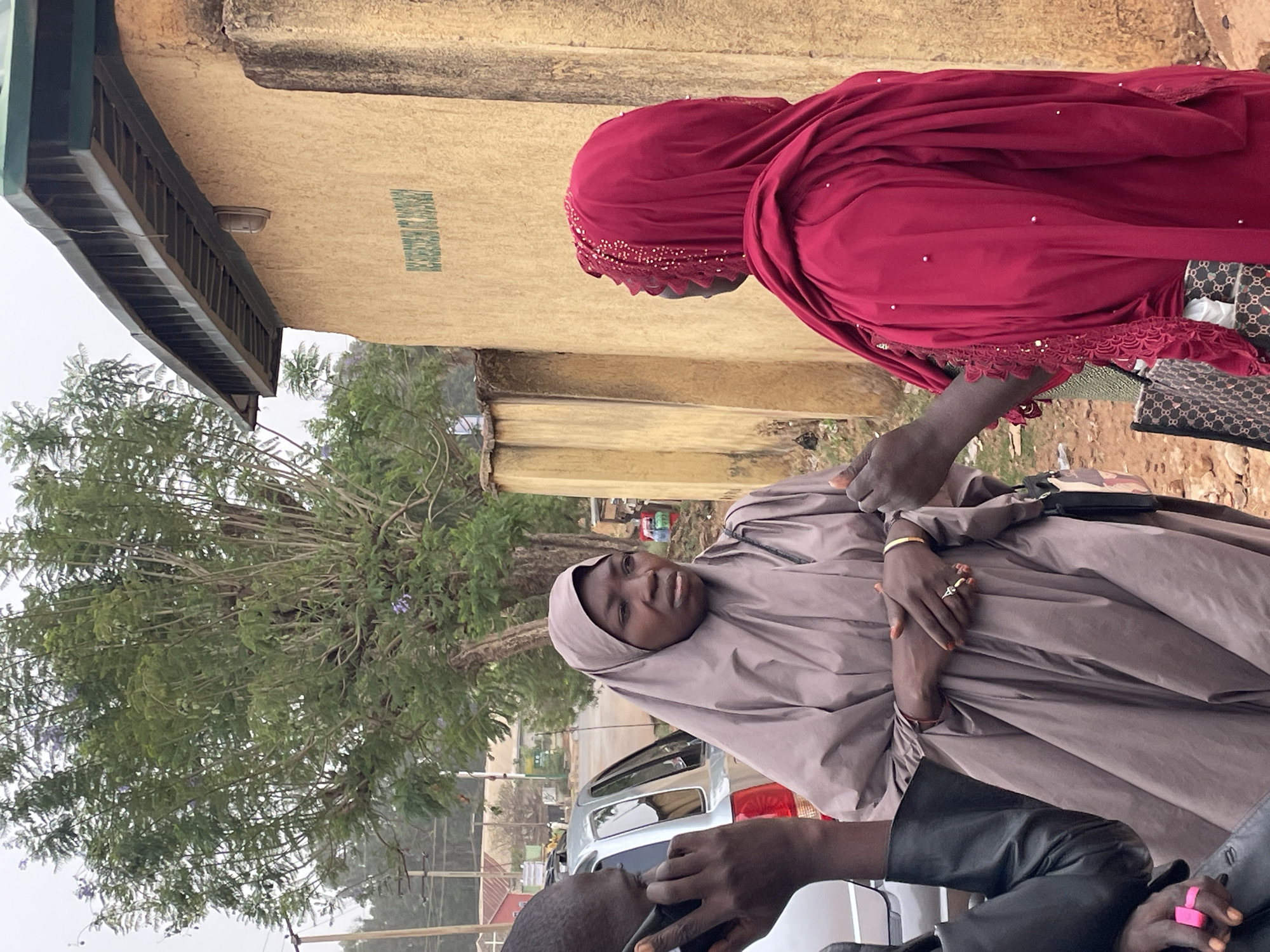 In a horizontally-oriented photograph, two women are prominently featured in the foreground, both wearing traditional Muslim attire. The woman on the left is dressed in a light brown hijab and robe, her hands clasped in front of her, adorned with rings. She is looking directly at the camera. The woman on the right is attired in a deep red hijab, with decorative embellishments along the edges. She appears to be interacting with the first woman.
Several other individuals are partially visible in the foreground. One individual is on the left using a mobile phone. Another individual wearing a black leather jacket is partially visible on the bottom right.
Behind them, a silver car is parked. A large tree with dense foliage occupies the left side of the background. Behind the women and tree, there is a pale yellow building with a green sign.
The setting appears to be outdoors, possibly a street or public area in the city of Jos, Nigeria.