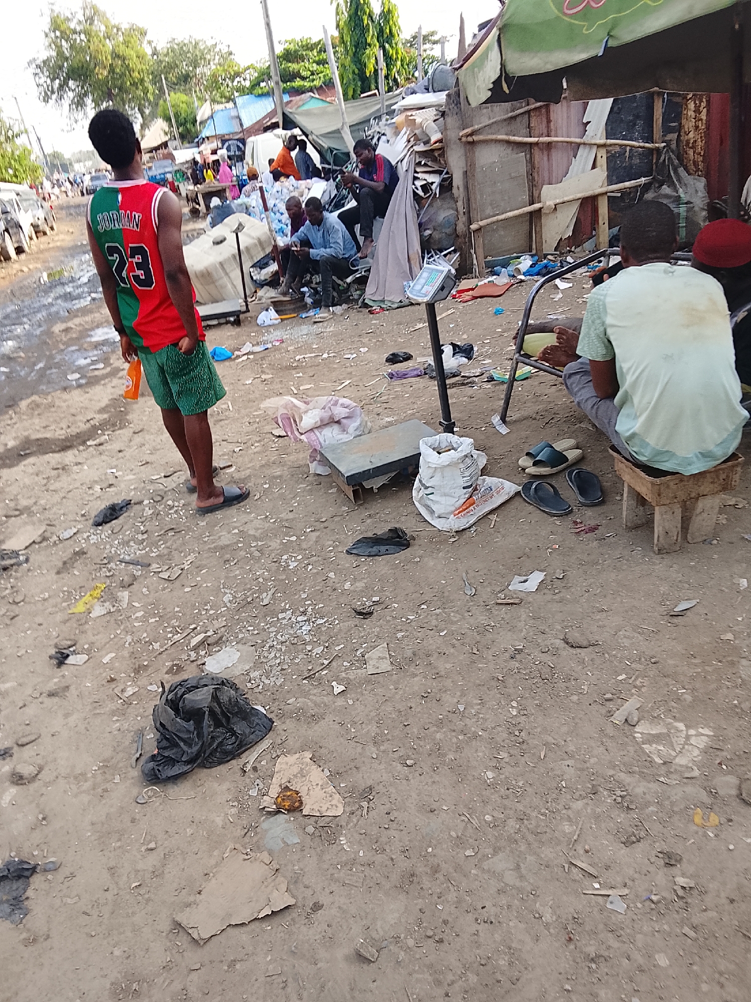 The image depicts a scene in an open, possibly makeshift market area in Abuja, Nigeria. The ground is dusty and littered with debris, including paper, plastic, and discarded items. Several people are present, engaged in various activities. One person in a "Jordan 23" jersey and green shorts stands facing the market. Other people are seated or gathered around what appears to be a weighing scale. A rudimentary structure made of scrap materials serves as a backdrop for some individuals. A bag and a piece of metal are next to the weighing scale. The general atmosphere suggests a low-income, informal trading environment.