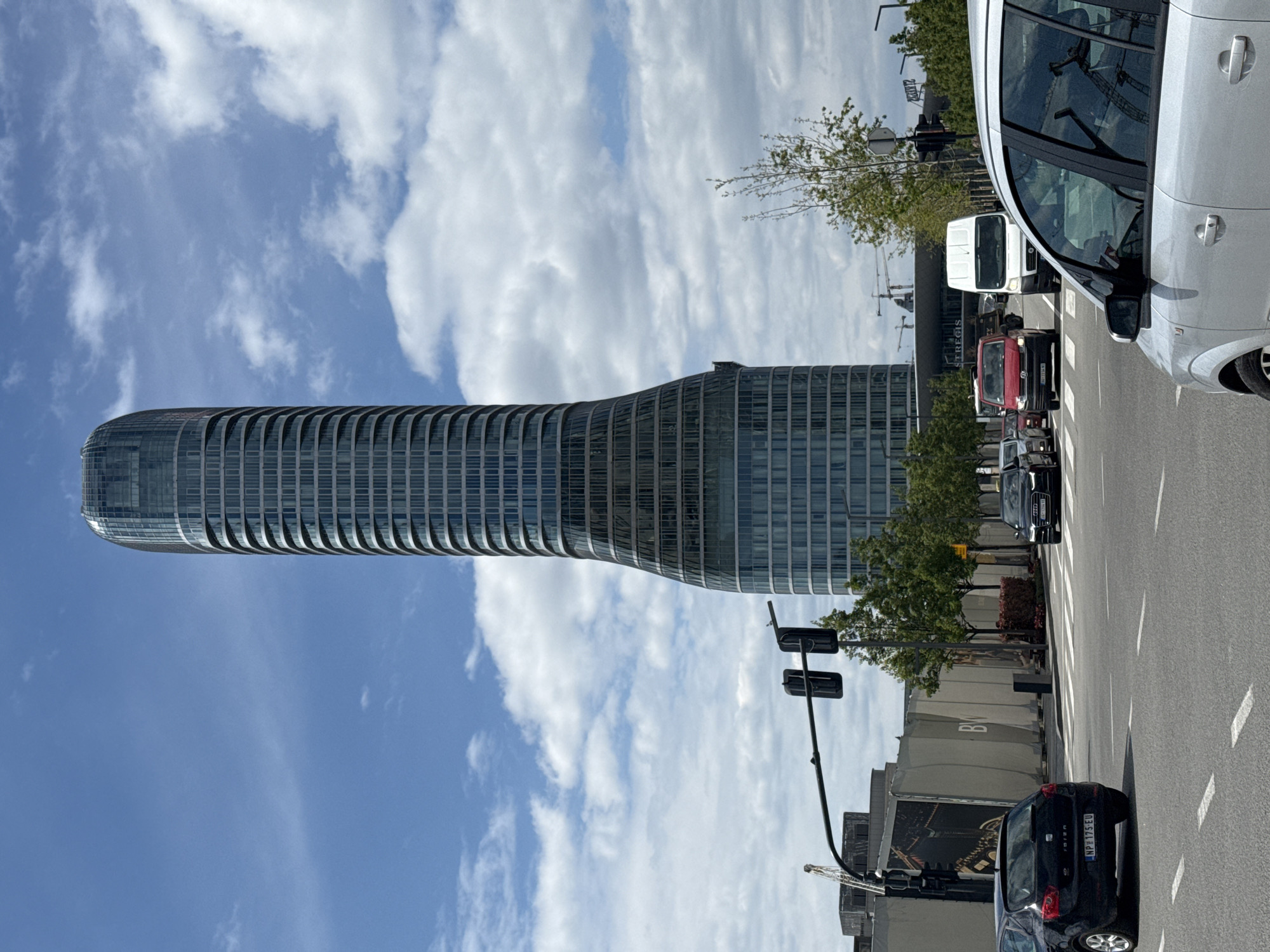 An upward-angled shot captures a vibrant daytime street scene in Belgrade, Serbia, dominated by a distinctive, modern skyscraper. The building, constructed from glass and horizontal white or light-grey panels, features a unique cylindrical form that tapers inward at its mid-section before widening again towards the base. Its reflective facade mirrors the bright blue sky and scattered white cumulus clouds.
In the foreground, a multi-lane asphalt road is visible, marked with white dashed lines. Several vehicles are present, indicating active city traffic. On the right, the front-right portion of a white car is prominent, showing its windshield and side mirror. Further down the street, a white delivery van and a red car are visible, with a black hatchback (license plate "NP 173EU") in the nearest lane.
Along the roadside, green trees with relatively sparse foliage line a paved area. Street infrastructure, including traffic lights and streetlights, is also visible. In the background, behind the trees and across the street, a construction site is apparent, marked by visible cranes and temporary barriers that display the letters "BW." Other building signage is partially visible, including what appears to be "TREC" and "ZARE" on different structures. The bright, direct light suggests it is late morning or early afternoon, with clear weather under a partly cloudy sky.