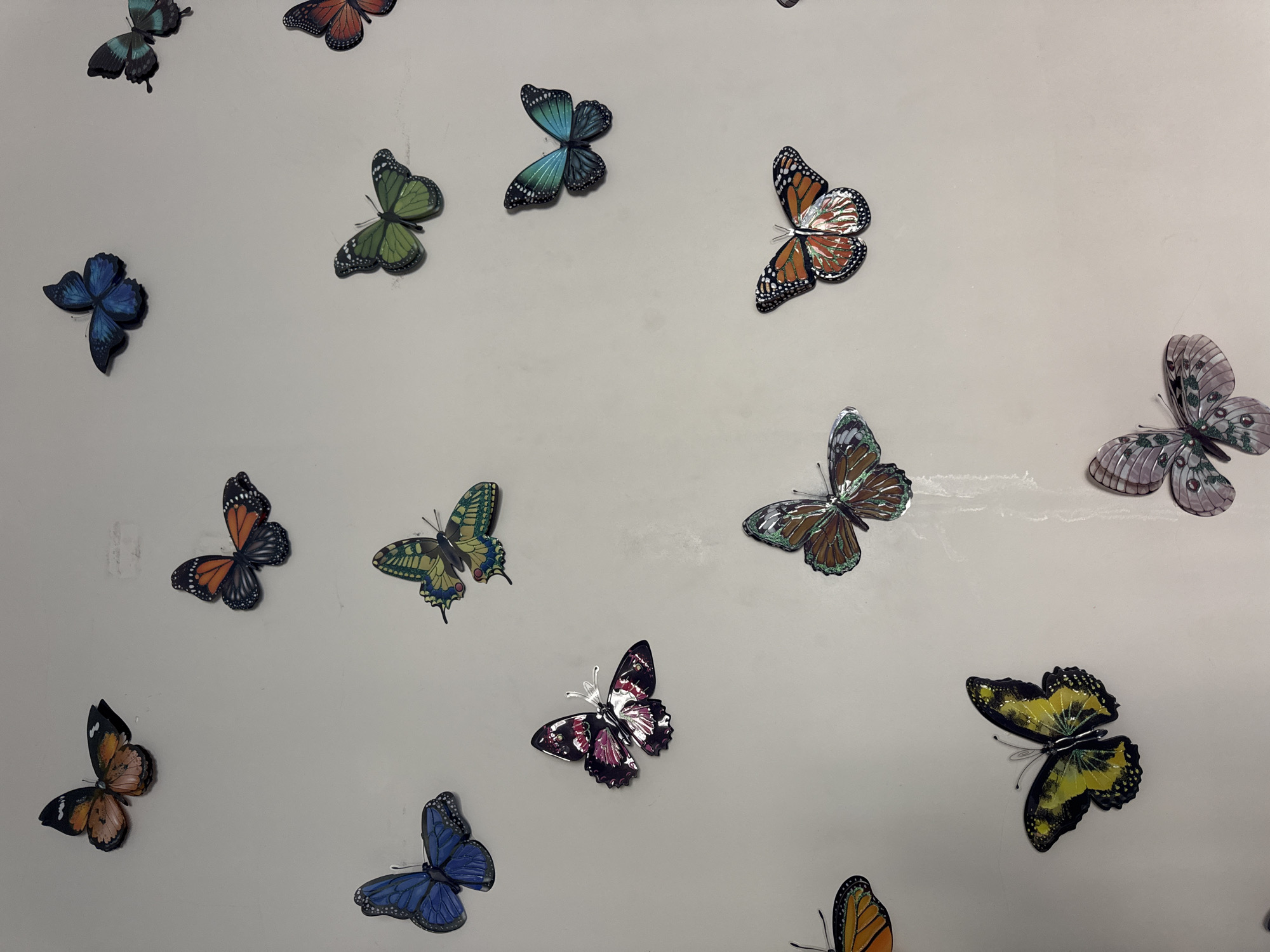 The image shows a collection of decorative butterfly ornaments adhered to a light gray surface. The butterflies vary in color, size, and pattern, with shades of blue, green, orange, brown, pink, and yellow represented. Some butterflies have visible antenna wire attachments. The composition of the butterflies appears random and scattered across the surface. There is no indication of the location of the image being in Kuta, Indonesia.
