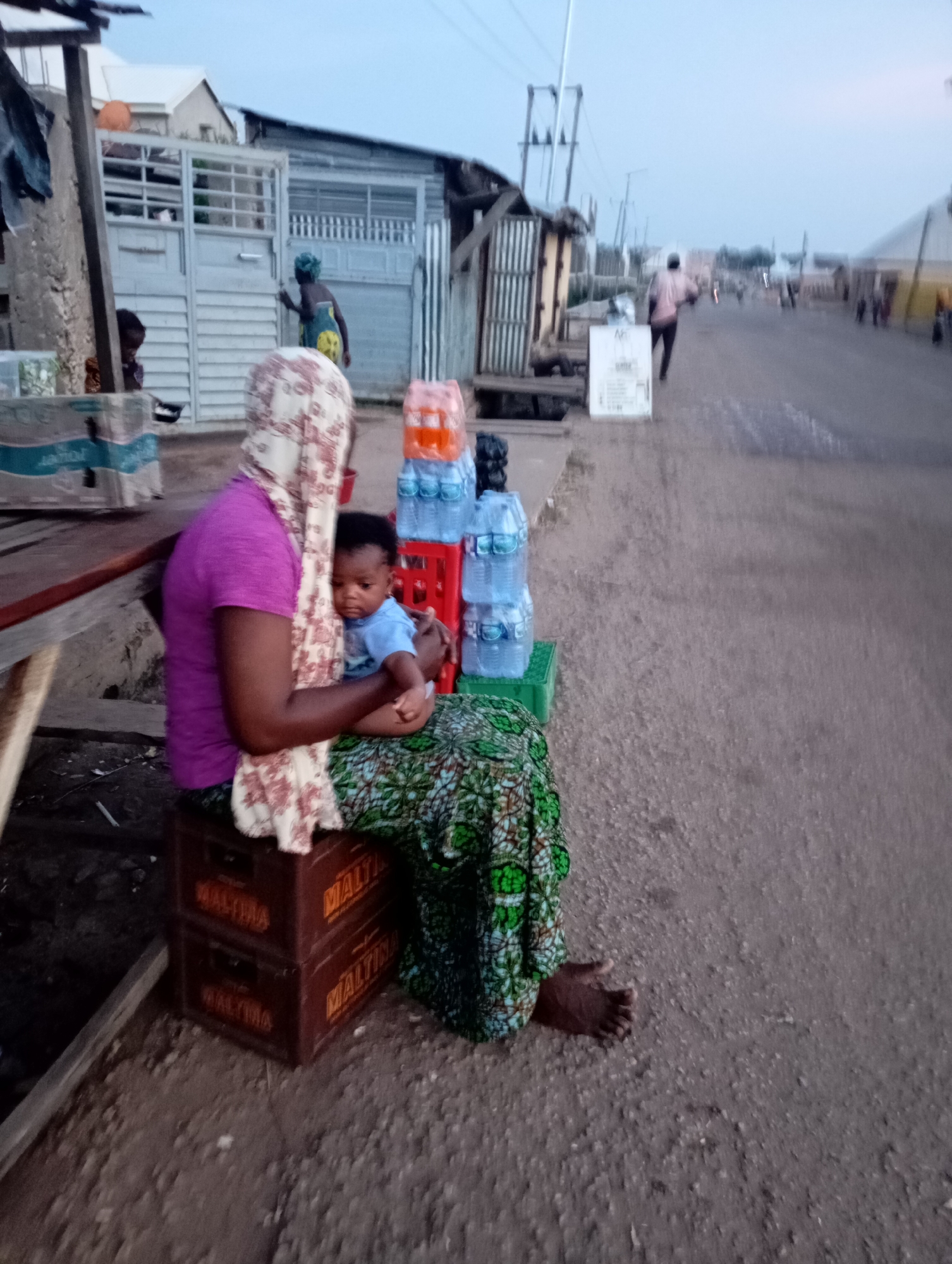 In Shendam, Nigeria, at dusk, a woman wearing a purple shirt and a patterned headscarf sits on two brown crates of maltina. She holds a baby in a blue shirt on her lap. To her right, stacks of plastic bottled water in red and green crates are visible. The background shows a street scene with buildings made of corrugated metal and concrete, a fence, and people walking in the distance. The ground is a dirt road.