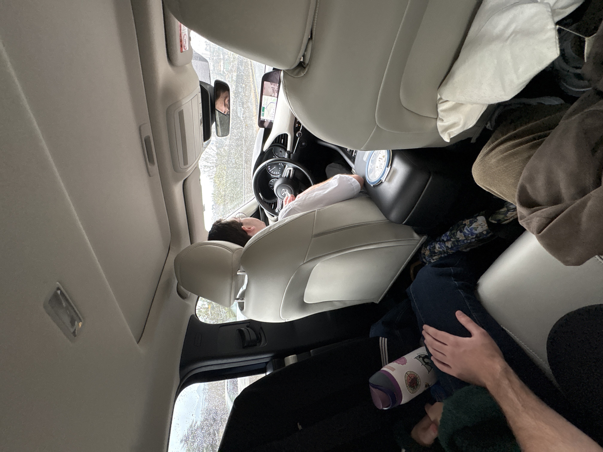 A car interior is depicted with a person in the driver's seat and individuals in the back. The driver is reclining, eyes closed, and resting their head against the headrest, wearing a white shirt. The rearview mirror reflects a person's face in the backseat. In the backseat, an individual is seated with their arm resting on their leg, wearing a dark green sweater. A patterned water bottle is visible near their leg. A white pillow or blanket is near the front passenger seat, and a brown garment is draped behind it. The car has light-colored upholstery and a sunroof. The exterior environment is visible through the car windows, with hints of foliage and buildings. The location is not directly identified as Santiago Yancuitlalpan, Mexico, in the image.