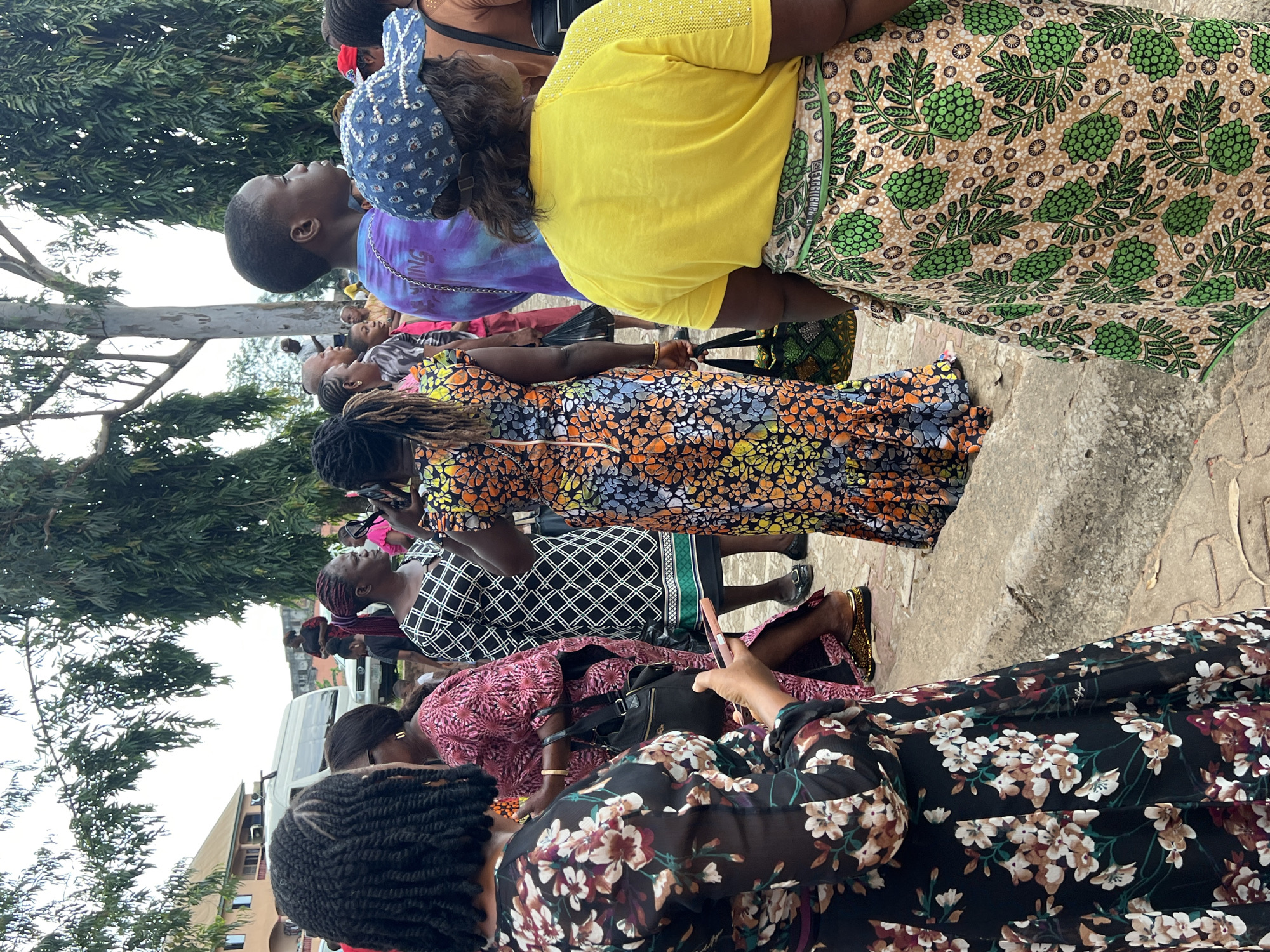 The image shows a group of people gathered outdoors in Nnewi, Nigeria. In the foreground, a woman with braided hair is looking at her phone. To her right, a woman in a pink patterned dress stands, holding a black handbag. Further to the right, a woman in a black and white geometric dress and a woman in a colorful floral dress are visible. In the background, a person in a tie-dye shirt is looking to the right, and another person is wearing a blue patterned cap. Several trees are present in the background, along with a building and a white van. The ground is paved with interlocking stones. The overall scene appears to be a public gathering or event.