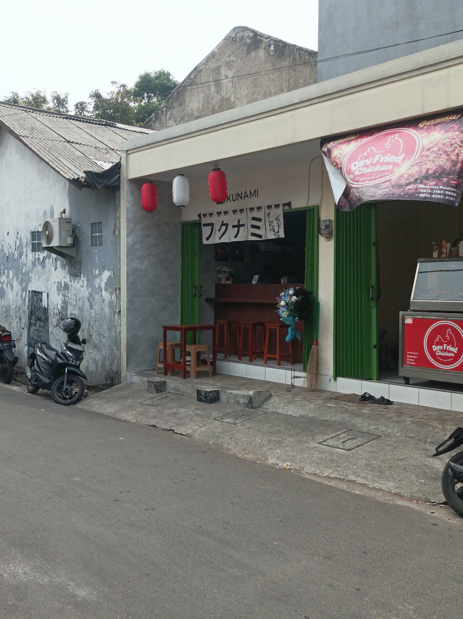 The image depicts the exterior of a small restaurant named "Dev Fried Chicken" with Japanese signage above the entrance. The restaurant is situated in a city, likely West Jakarta, Indonesia, as suggested by the architecture and signage. The building itself is a modest structure with a light-colored facade and green bi-fold doors that are partially open, revealing the interior.
In front of the restaurant, there is a street with a gray asphalt surface. On the left side of the street, a dark-colored motorcycle is parked. The building to the left of the restaurant is older and weathered, with peeling paint and a corrugated metal roof. An air conditioning unit is mounted on its wall.
Above the entrance of "Dev Fried Chicken," several red and white paper lanterns are hung. A banner displaying the restaurant's name and a slogan, "Crispy and juicy!," is partially visible on the right side of the building. Inside the restaurant, through the open doors, one can see a counter with stools and what appears to be a display case for food. A broom and a decorative flower arrangement are placed near the entrance. The ground in front of the restaurant is paved with concrete.
The overall scene suggests a casual dining establishment in a bustling urban environment. The weather appears to be overcast, with diffused light.