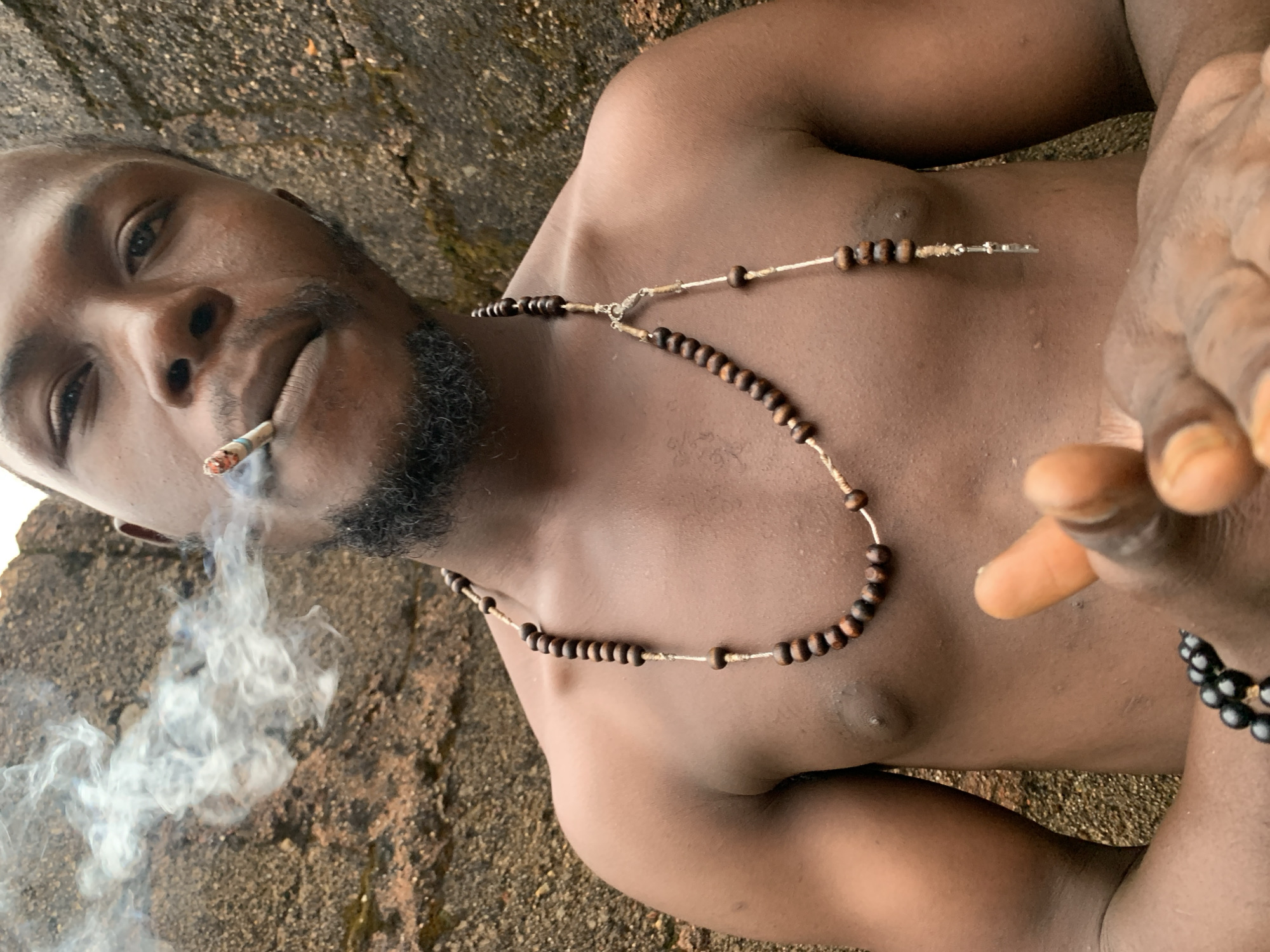 The image shows a close-up, chest-up view of a dark-skinned man smoking. He is shirtless, wearing a necklace made of brown beads and a second beaded bracelet on his left wrist. He has a goatee and is exhaling smoke. He is positioned in front of a rough, textured rock surface. The lighting suggests it is daytime. The background is a reddish-brown rocky surface, possibly a natural formation or a wall. The man's skin appears slightly moist, suggesting either sweat or humidity. There are no discernible urban features or landmarks that would place this image specifically in Jos, Nigeria, or any other city. The setting appears to be natural or possibly an outdoor urban area with exposed rock.