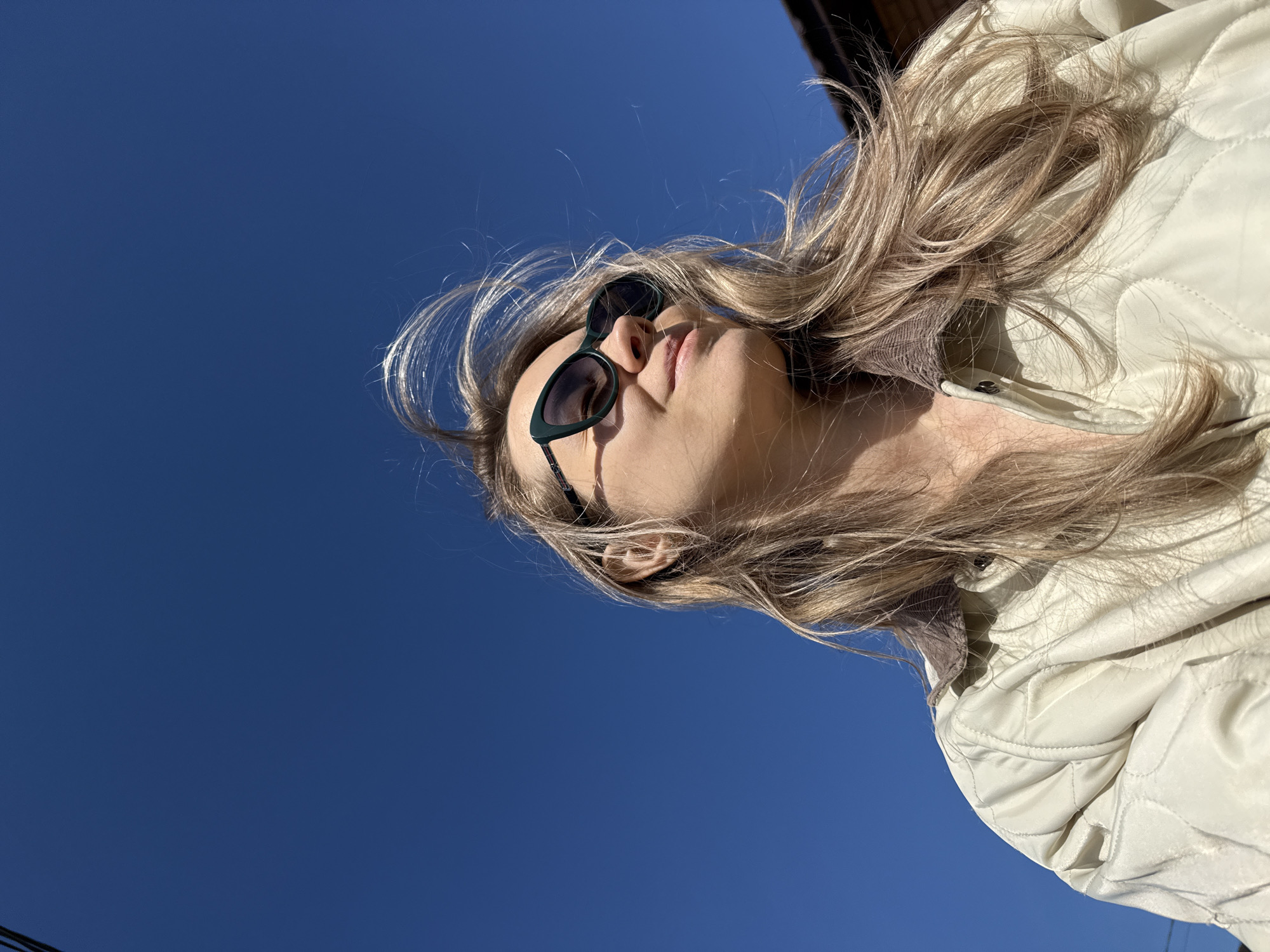 A low-angle shot of a person in Chaikovsky, Russia. The person, wearing dark sunglasses and a light-colored quilted jacket, is oriented at an angle against a clear, deep blue sky. The background shows a corner of a dark-colored structure. The hair is light-colored and windblown.