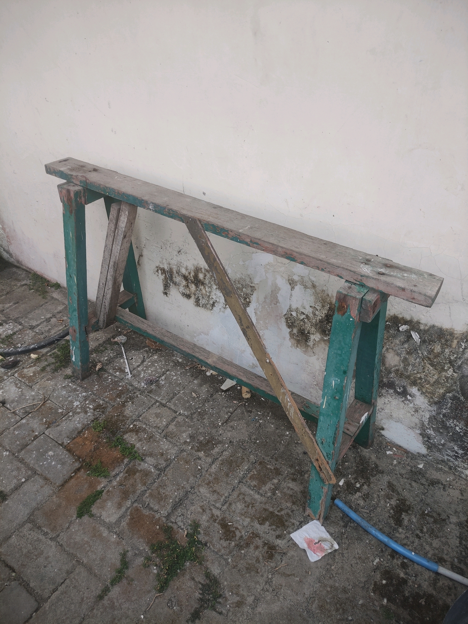 An aged, green-painted wooden sawhorse stands against a weathered, off-white wall. The sawhorse, constructed from rough lumber, appears worn and weathered, with visible nail heads and some peeling paint. It is positioned on a paved ground surface composed of interlocking grey bricks, some of which are cracked or have patches of dirt and sparse green vegetation growing between them. The wall behind the sawhorse exhibits signs of age and dirt, with dark, splotchy discoloration.
To the left of the sawhorse, a black hose lies on the ground. On the ground near the front right leg of the sawhorse, there is a crumpled white tissue with a pink stain and a blue plastic tube. The scene is outdoors, likely in a yard or construction area. The overall impression is one of disuse and the passage of time. The location is identified as Ngasem, Indonesia.