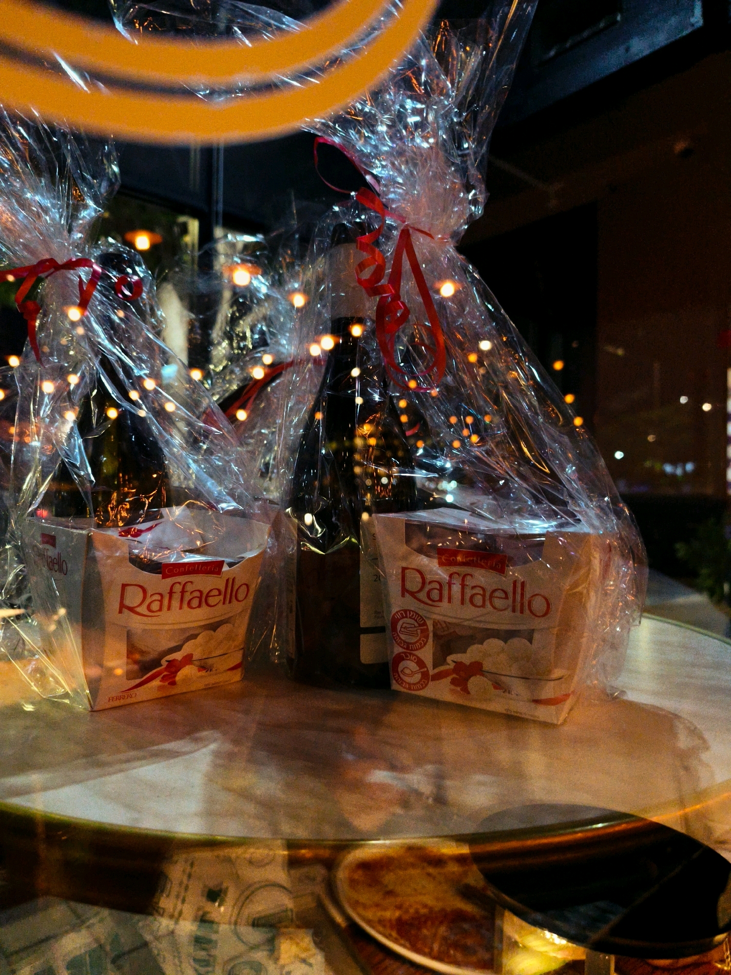 The image shows two gift baskets containing RaffaELLO chocolates and bottles of wine, wrapped in clear cellophane and tied with red ribbons. The baskets are placed on a reflective table, likely outdoors, as indicated by the bokeh effect of lights in the background, which suggests a nighttime setting with ambient lighting. One basket is more prominent in the foreground, displaying the RaffaELLO logo clearly. The background is dark and blurred, with streaks of orange and white lights, suggesting an urban environment. The table surface reflects the objects and lights, adding depth to the scene. The overall impression is that of a festive or celebratory presentation of gifts. The specific location in Be'er Sheva, Israel is not discernible from the image itself, but the presence of Hebrew text on the RaffaELLO packaging hints at the region.