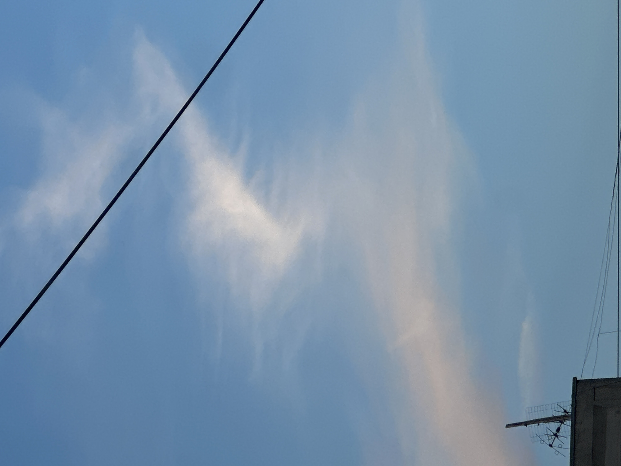This media file captures a low-angle shot of a light blue sky with wispy white and pinkish clouds. A dark power line runs diagonally from the top left towards the center of the frame. In the lower right corner, the top of a concrete structure is visible, with an antenna attached to it. The antenna has a grid-like reflector and several dipole elements. The scene suggests it might be late afternoon or early evening due to the warm tones in some of the clouds. The weather appears clear with scattered clouds. The presence of the power line and antenna suggests an urban or semi-urban environment. There is no visible text in the media file.