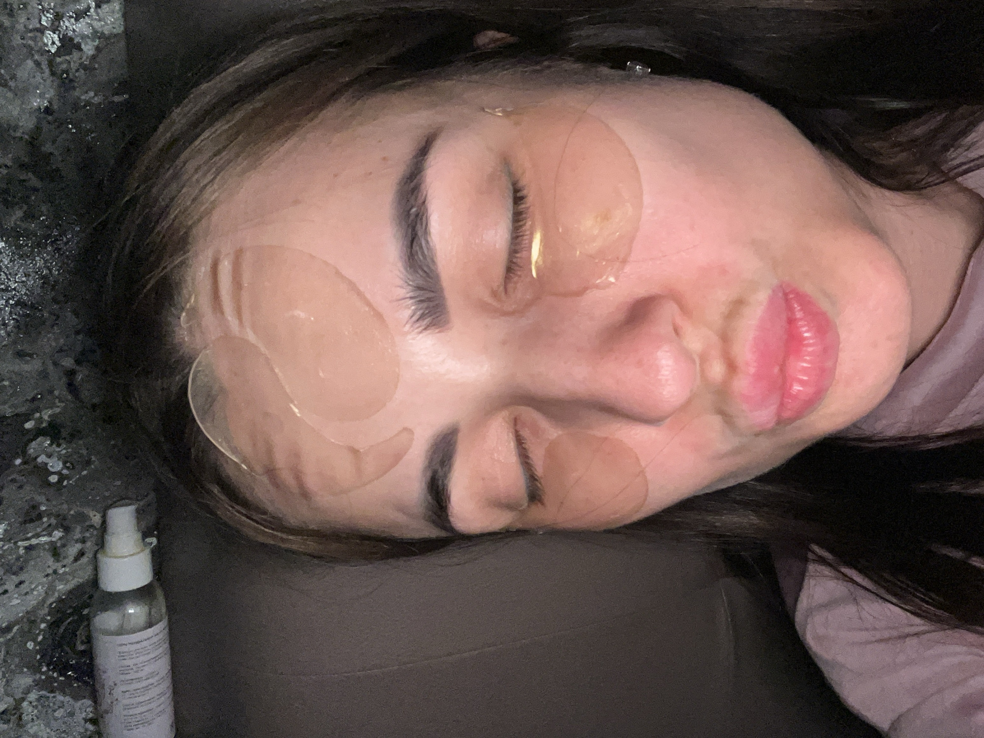 A close-up, slightly angled shot depicts a woman with dark hair lying on a dark grey surface. Her eyes are closed, and her face is adorned with several translucent gel patches. To the left of the woman, on a speckled surface, lies a small, clear spray bottle. The location is Unknown, Unknown.
