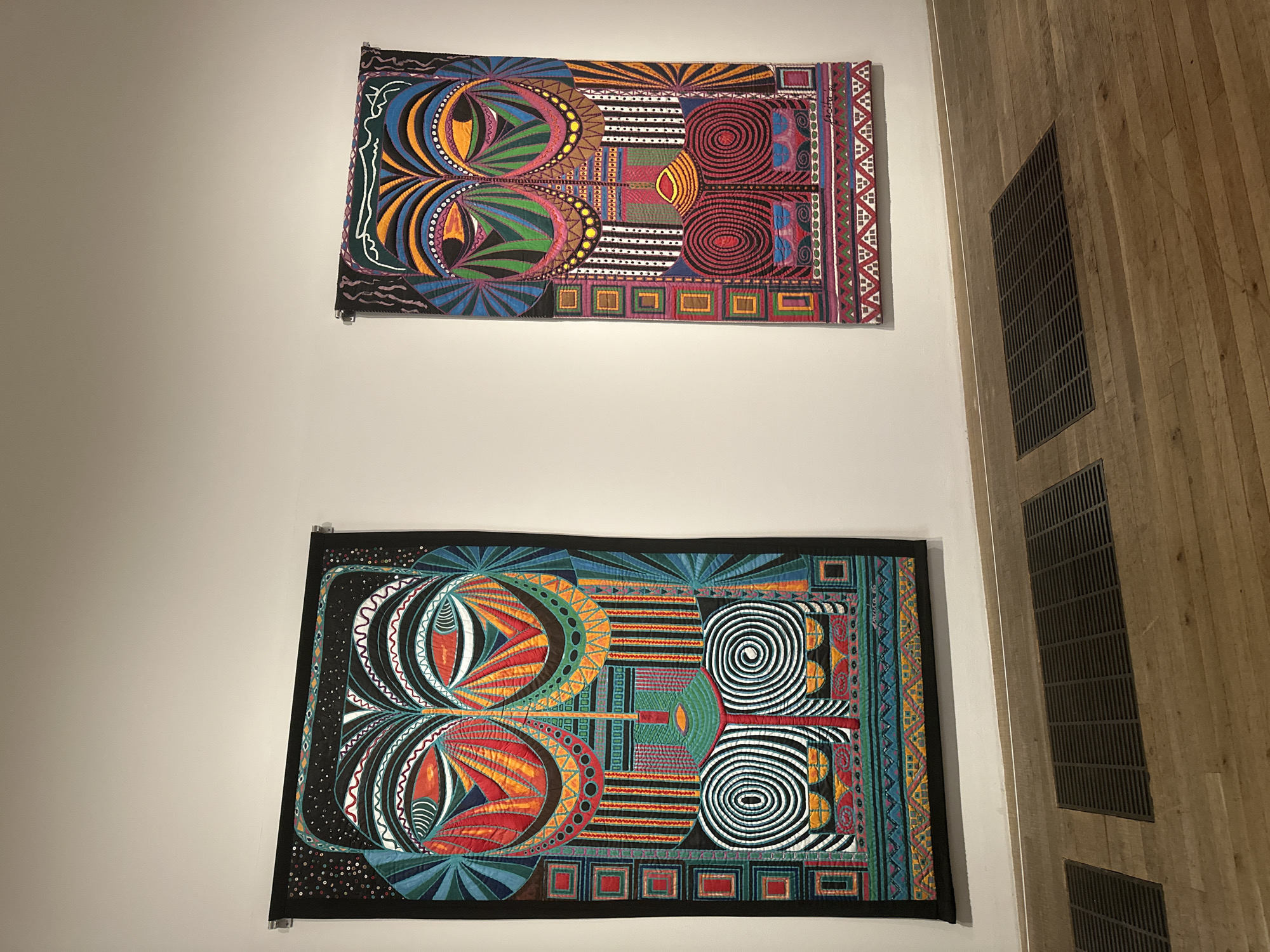 Two vibrant, intricately quilted artworks hang on a white wall, depicting stylized masks with large, colorful eyes and geometric patterns. The artwork on the left is larger and predominantly features black, red, orange, and teal. The artwork on the right is slightly smaller and incorporates more purple and magenta into its color scheme. Both pieces have black borders and appear to be in a gallery setting, indicated by the clean white walls, wooden floor, and discreet ventilation grates. The lighting suggests an indoor exhibition space. The scene captures a moment of quiet observation, with no people directly visible in the frame.