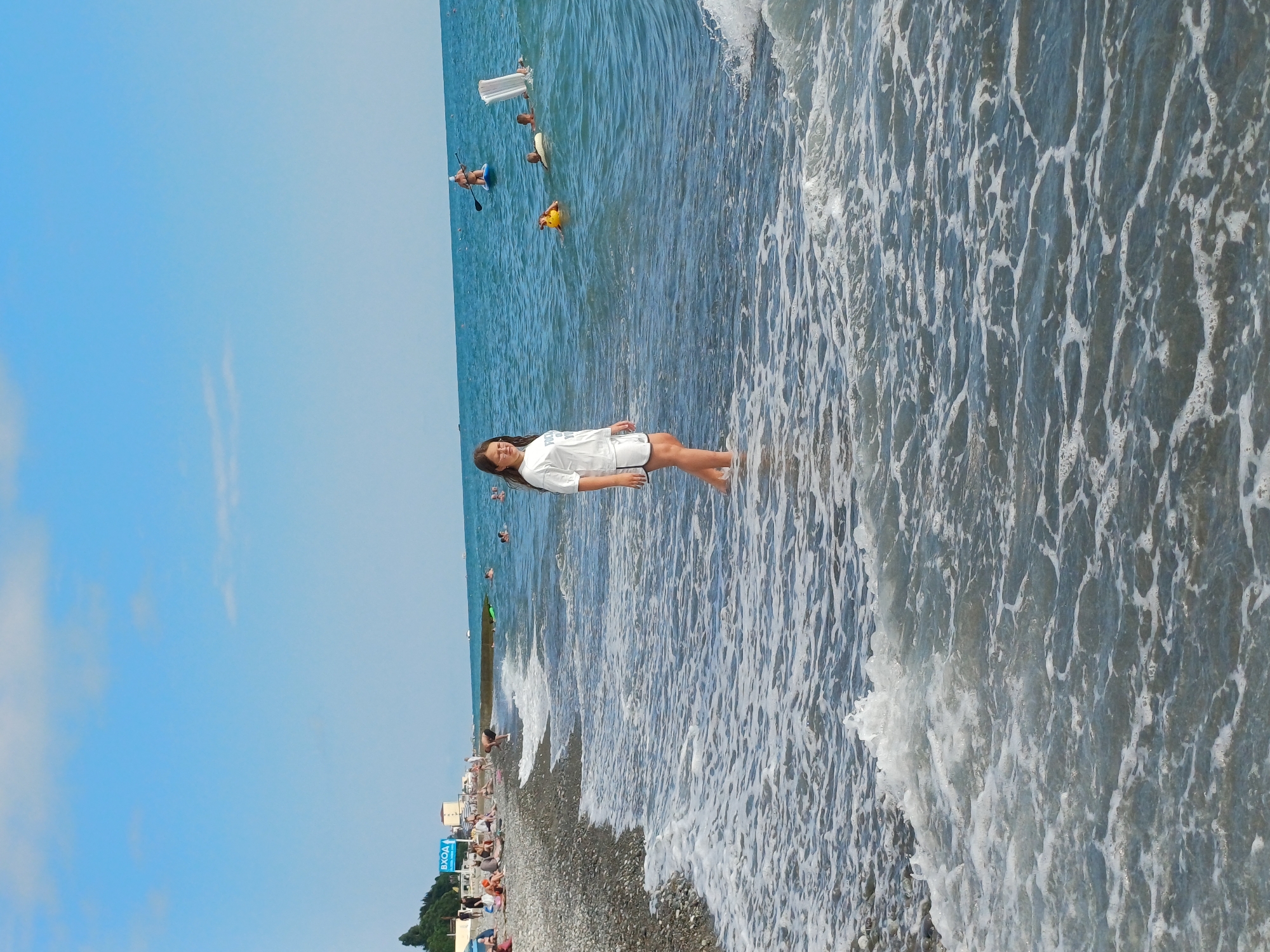 The image shows a sunny day at a pebble beach in Sochi, Russia. The focus is on a young woman standing in the shallow water at the edge of the sea, wearing a white t-shirt and dark shorts. Waves are gently lapping around her feet. In the background, the blue ocean stretches out, with several people enjoying various water activities. Some are on paddleboards, one person is on a floating mattress, and others appear to be swimming or wading. Further in the distance, the coastline is visible with some buildings and trees, and a line of people are relaxing on the beach. The sky is a clear, bright blue with a few wispy clouds. A blue sign with white Cyrillic text reading "ВХОД" (which translates to "ENTRANCE") is visible on the beach. The overall mood is relaxed and summery.