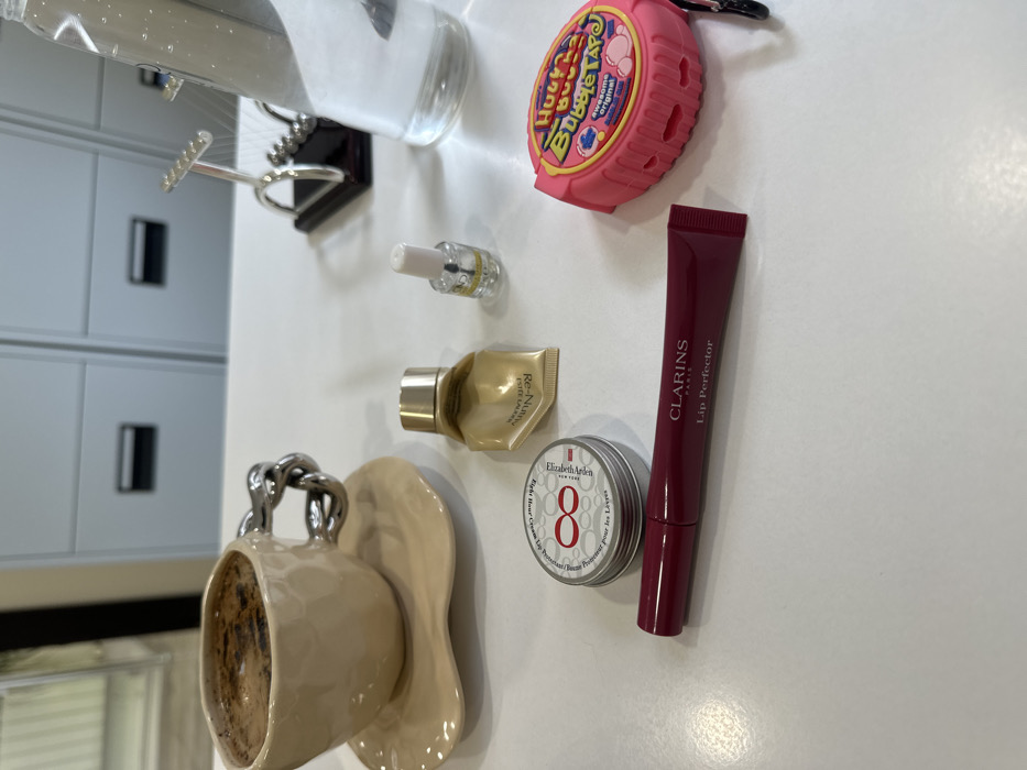 The image features several objects on a white surface. Starting from the top, there's a bottle, followed by "Hubba Bubba Bubble Tape" in a pink container. There's also a small bottle of nail polish and a travel-sized "Re-Nutriv" moisturizer. Below those, there is a metal tin of "Elizabeth Arden Eight Hour Cream," and a "Clarins Paris Lip Perfector" in a dark red tube. A decorative beige cup and saucer with a chain handle is displayed on the left. A white storage cabinet with a black handle is in the background.