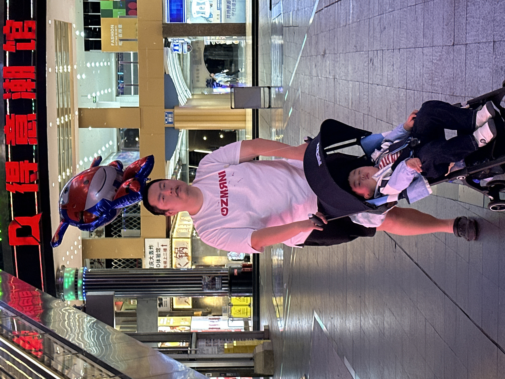 A medium shot captures a man pushing a child in a stroller on a pedestrian walkway in Yuzhong District, Chongqing, China. The walkway is constructed of large, light-gray, rectangular pavers. The man is fair-skinned and wears a white, short-sleeved t-shirt with red lettering, dark shorts, and dark shoes. The child in the stroller wears a light-colored jacket and dark pants, and is wearing white shoes. To the left of the man, a red, white, and blue balloon in the shape of a cartoon character is visible.
The background is filled with businesses and advertisements. A red neon sign with Chinese characters is prominently displayed. There's also an escalator to the left, partially visible. Several store signs are present. One reads "FASHION EATING IN DEYI 吃在得意". Another reads "庆大轰炸 D体验馆一 电梯上二楼↑锅". The background also shows the architecture of surrounding buildings with lighted signs. The lighting suggests it is evening or night.