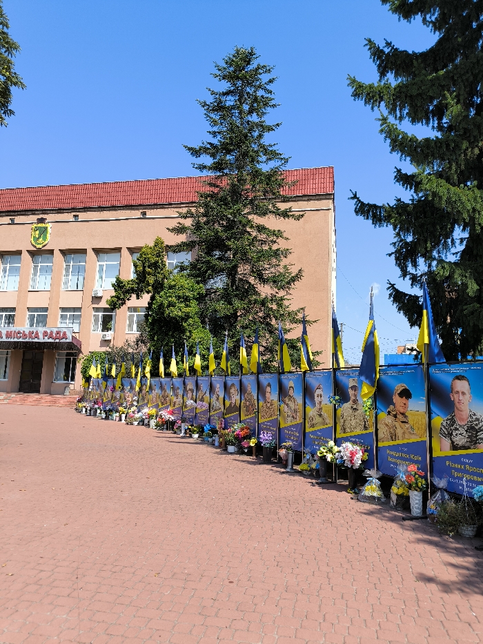 An outdoor scene in Illintsi, Ukraine, displays a memorial installation positioned along a paved area in front of a civic building identified as the "МІСЬКА РАДА" (City Council). The two-story, light-brown building features a red-tiled roof, multiple white-framed windows, and a yellow and green crest on its facade. A large evergreen tree stands centrally before the building, with additional trees framing the view. The memorial consists of a curved line of vertical banners, each approximately human height, displaying a portrait of an individual, primarily in military uniform, against a blue and yellow background with accompanying Ukrainian text. Small Ukrainian flags are mounted on poles at the top of each banner. At the base of each banner, on the red brick paving stones, numerous floral tributes, including bouquets of fresh and artificial flowers in various colors and potted plants, are arranged in dark containers. The sky is clear blue. No individuals are present in the image.