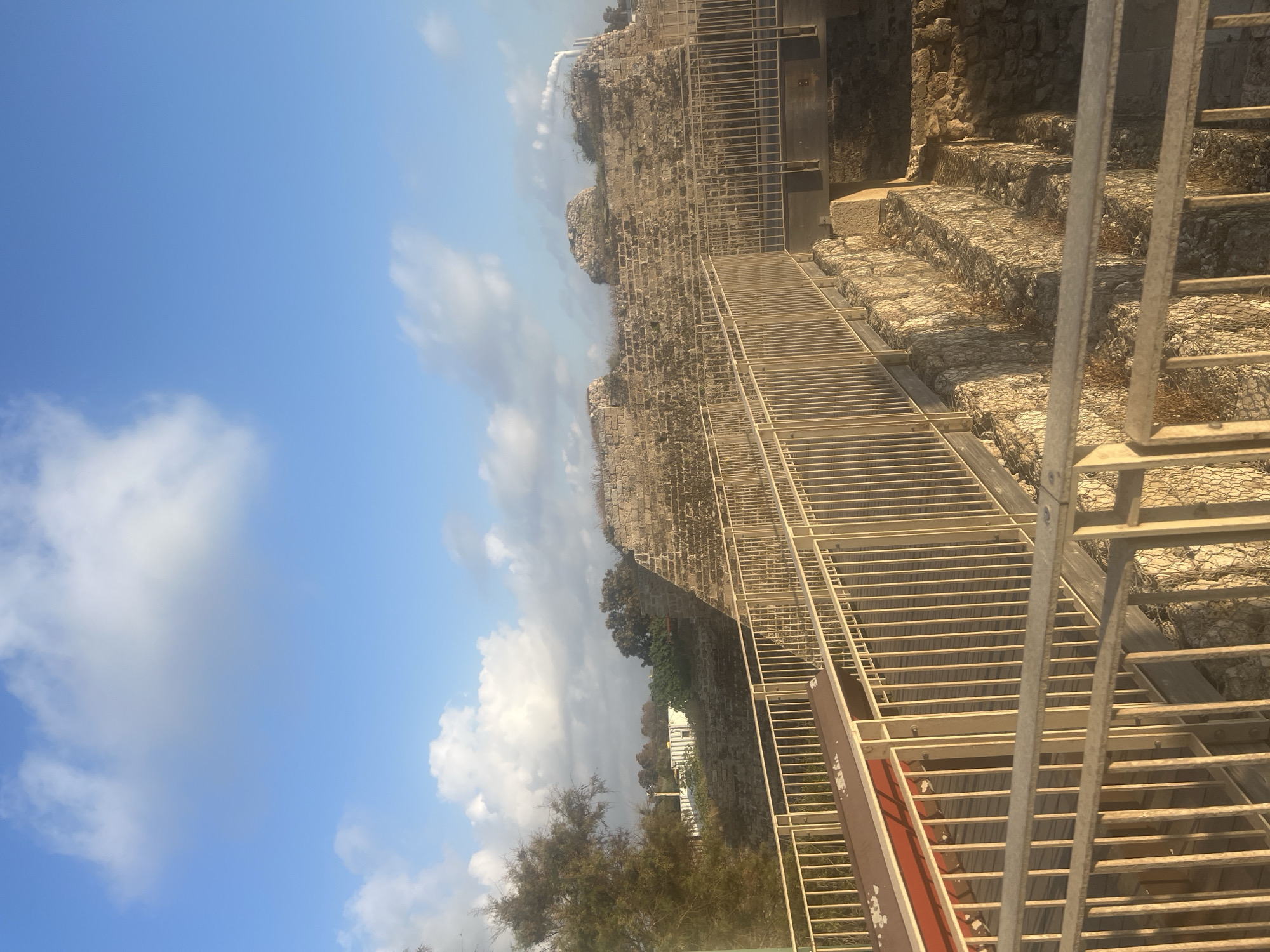 The image captures an archaeological site in the city of Hof HaCarmel Regional Council, Israel. The scene is dominated by ancient stone structures, likely remnants of a fortification or historical building, with visible brickwork and weathered stones. A modern walkway with a metal railing, possibly for tourists or researchers, traverses the foreground, offering a pathway over exposed excavation areas. These excavated sections reveal more stone foundations and structures, suggesting layers of history. The environment is characterized by a clear blue sky with scattered white clouds, and a few trees with sparse foliage are visible in the background, along with a distant industrial structure emitting smoke. The lighting suggests it might be late afternoon or early morning, with shadows cast on the structures. There are no people or animals present in the frame.