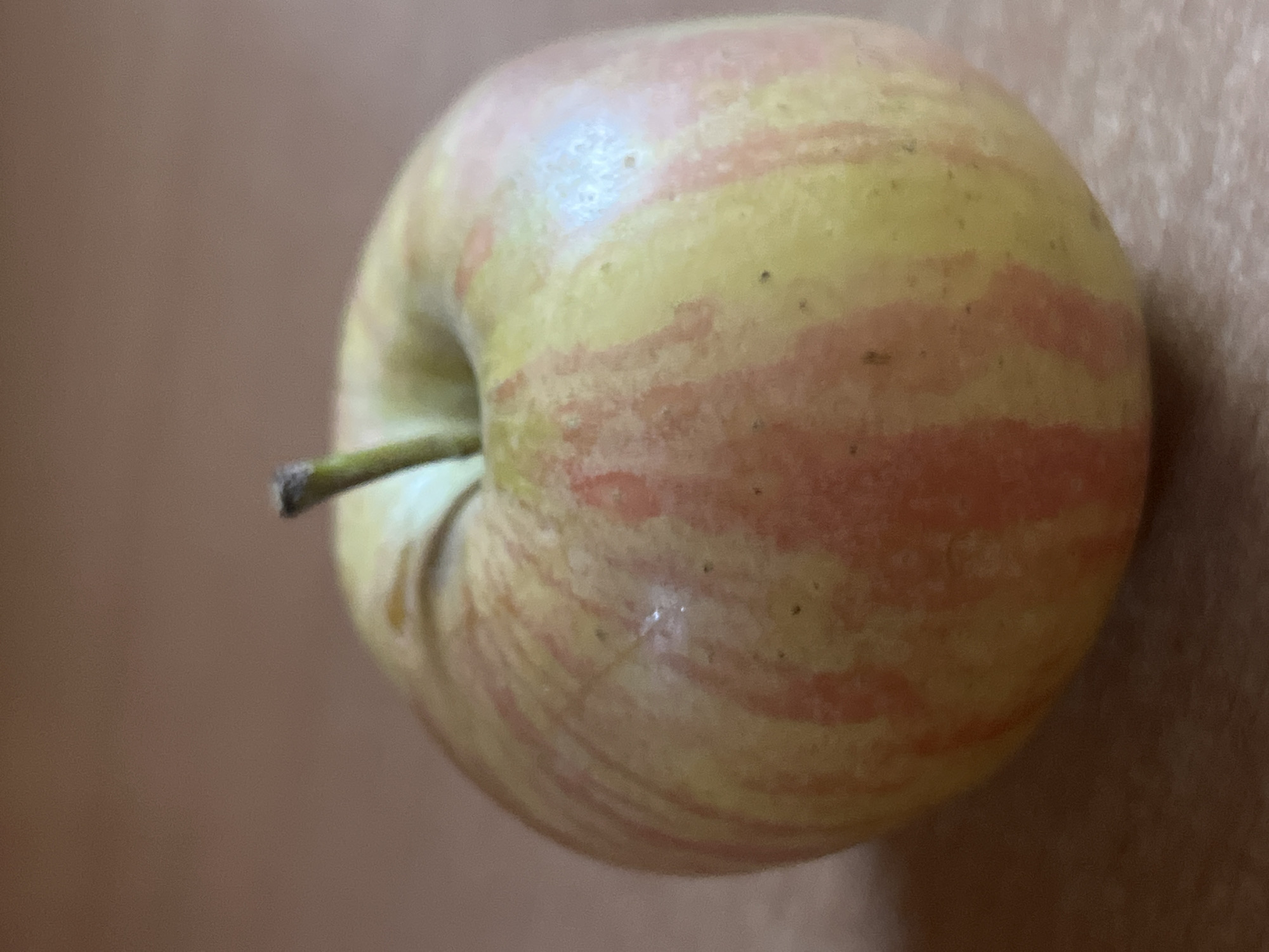 A single, small, ripe apple is depicted in a close-up, eye-level shot. The apple is primarily yellow with streaks of pink and red blush. Its stem, which is short and dark brown, is visible at the top center of the fruit. The apple is positioned on a flat, brown surface, possibly a table or countertop. The background is a blurred, muted brown color. There is no indication of any people, actions, or interactions. The image appears to be taken indoors, with soft lighting that highlights the apple's texture and color. The location of Iksha, Russia, is not determinable from the visual information presented in the image.
