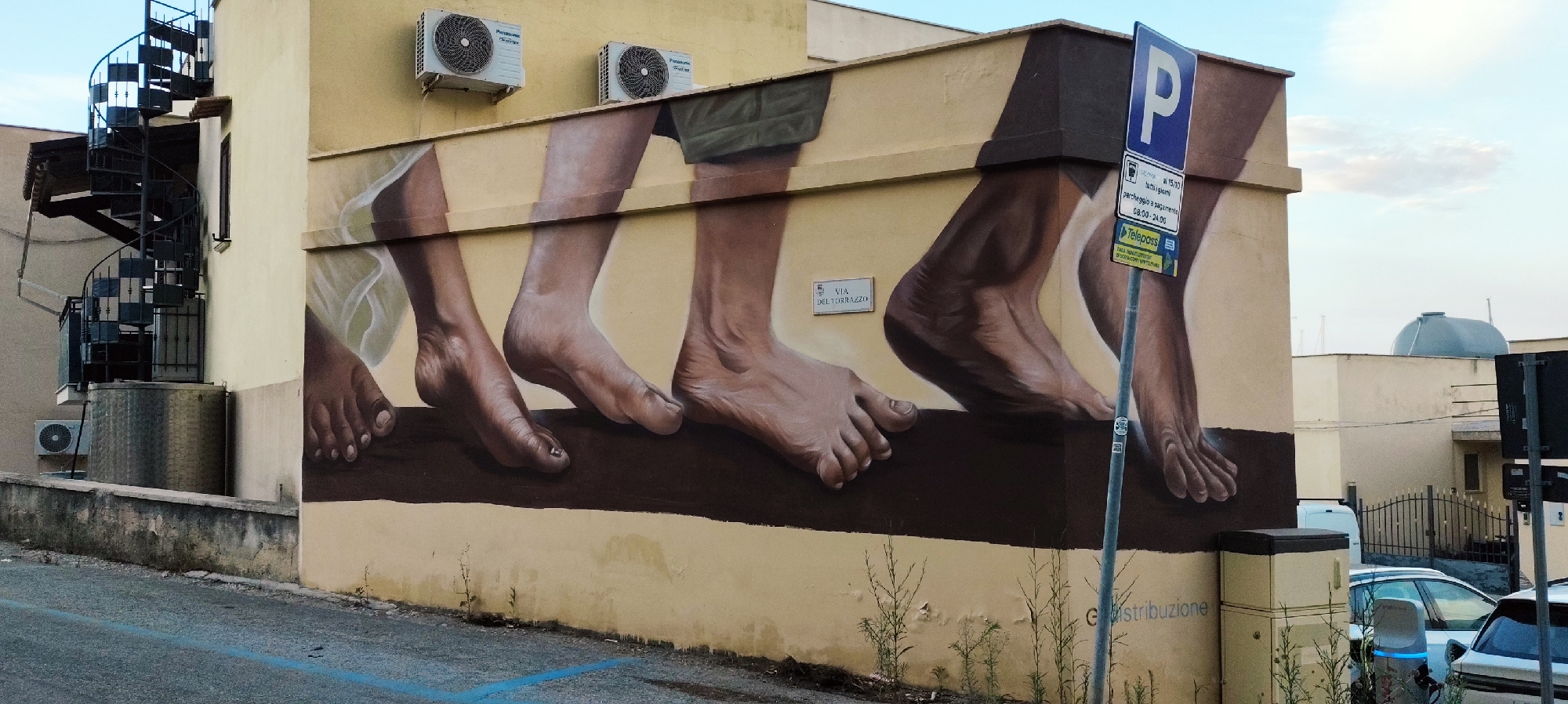 The image shows a mural painted on the side of a building in San Vito Lo Capo, Italy. The mural depicts several pairs of bare feet walking across a dark brown surface. The feet appear realistic and are painted with detail, showing toes and the soles of the feet. The background of the mural is a light yellow color, with a lighter shade behind the feet. Above the mural, on the yellow wall, are two air conditioning units.
To the left of the mural, a dark metal spiral staircase ascends the side of a building. Below the staircase, a large cylindrical metal tank is visible. A smaller air conditioning unit is mounted on the wall below the tank.
In front of the mural, a blue and white parking sign is mounted on a metal pole. The sign indicates parking is allowed at a cost of €1.50 all day, from 08:00 to 24:00, and also shows a Telepass logo. A street sign on the wall reads "VIA DEL TORRAZZO".
In the background, other buildings are visible, including one with a domed roof. A black metal fence separates a property from the street. Several vehicles are parked in the background, including a white car and a silver car. The sky is visible at the top of the image, with some clouds. The ground in the foreground is an asphalt road with a blue line painted on it. Some sparse green weeds are growing at the base of the mural.