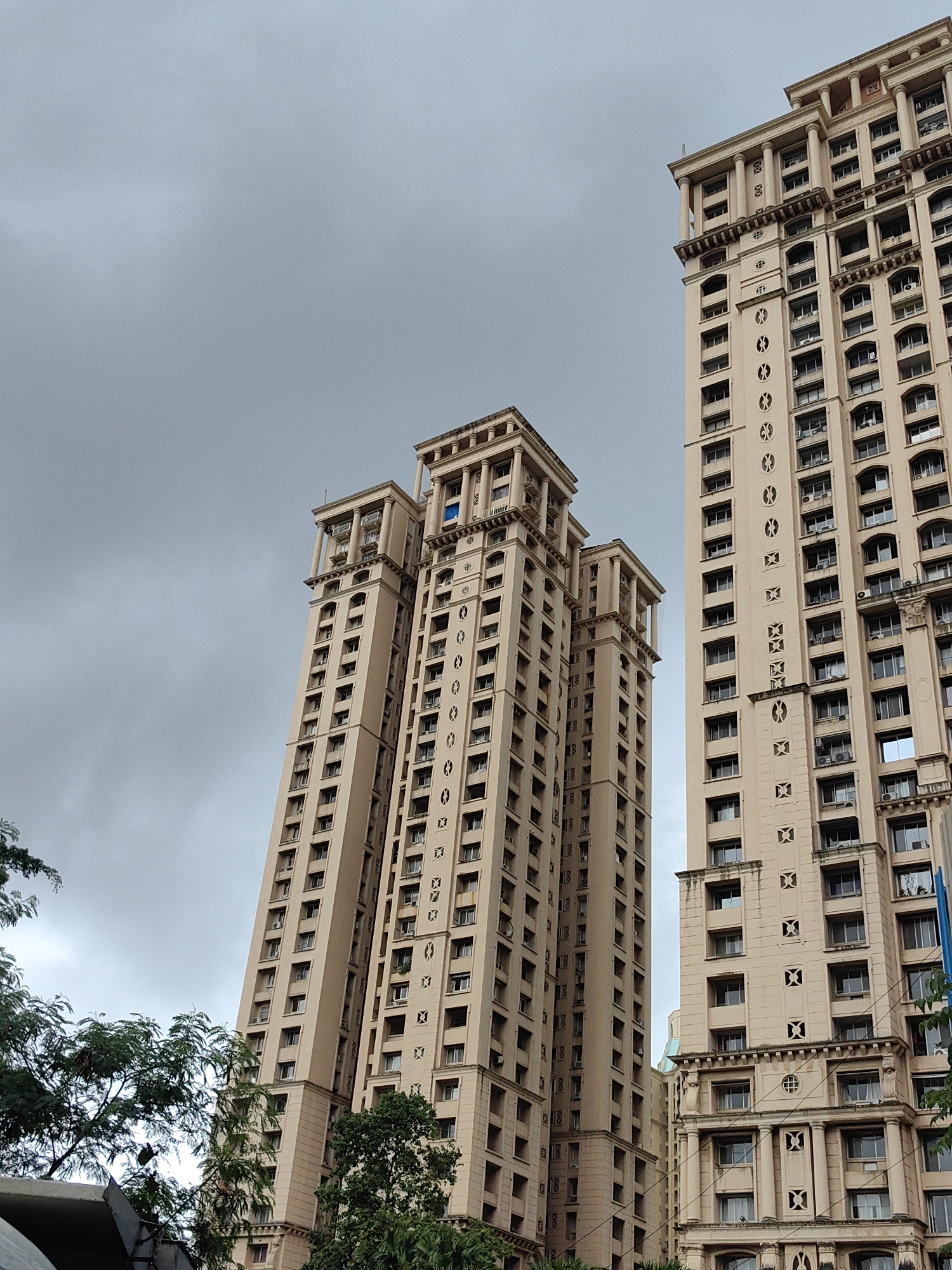 A low-angle, wide shot captures three tall, beige apartment buildings against a heavily overcast sky in Mumbai, India. The buildings, characterized by their ornate architectural details including columns and patterned facades, are arranged in a staggered formation, with the building on the right being the tallest and widest. The sky is a uniform, pale gray, suggesting a cloudy or pre-rainy day. Lush green trees are visible in the foreground on the left and partially obscuring the base of the leftmost building. The image appears to be a static architectural photograph taken from street level. There are no people or any observable action within the frame.
