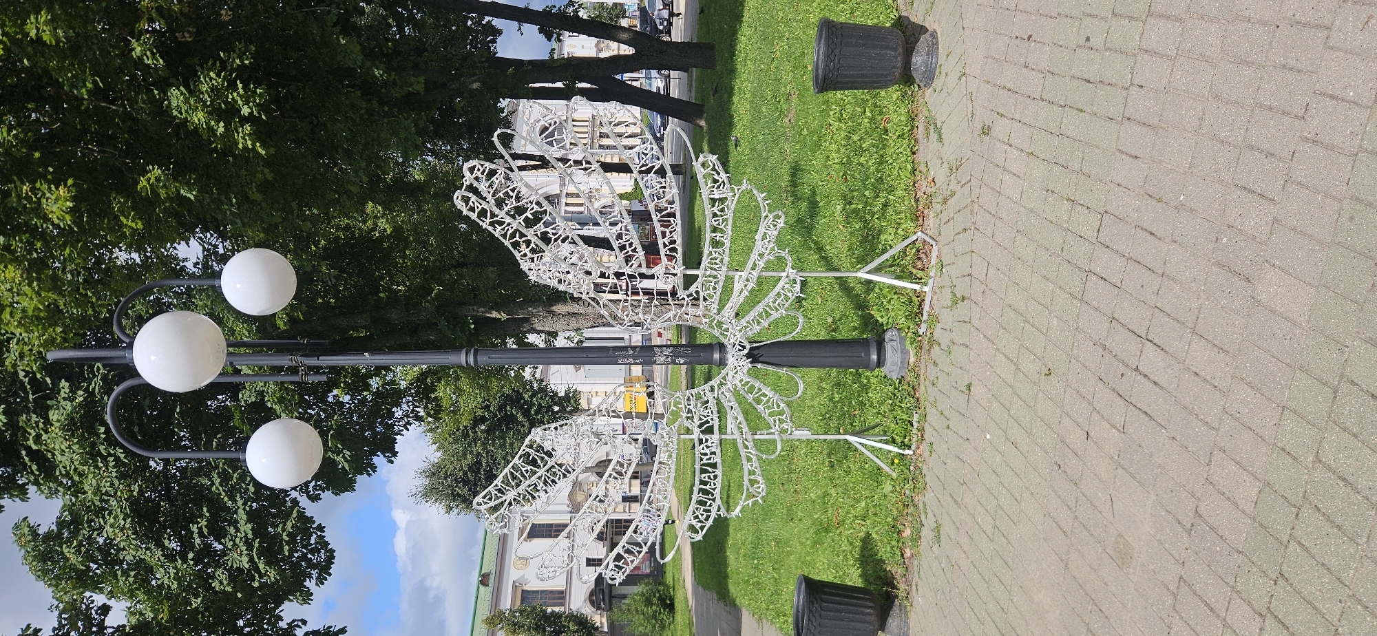 A black lamppost with three spherical white lights stands on a paved area in a park-like setting. Attached to the lamppost at its base is a large, white, wireframe sculpture shaped like a pair of open wings, likely made of lights. The sculpture is supported by metal stands. To the left and right of the lamppost and sculpture are two black, urn-shaped trash receptacles. The ground behind the sculpture is covered in green grass with some patchy areas and sparse weeds. In the background, there are several large, mature trees with green foliage. Beyond the trees, a white building with a green roof and arched windows is visible, along with other buildings and some parked cars. The sky is partly cloudy. The image appears to have been taken during daylight. The scene is located on Frunze Street in Vitebsk, Belarus, near house number 11, which is the building in the background.