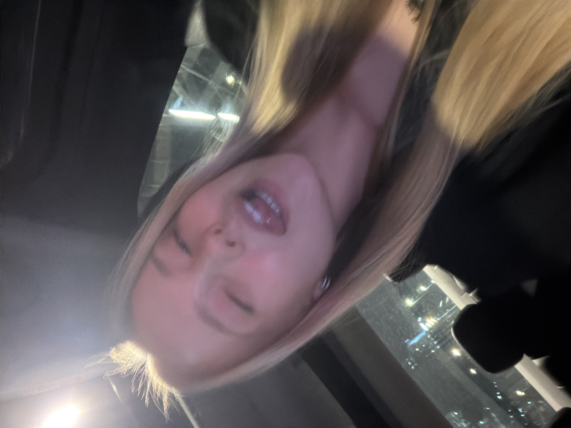 A close-up, angled photograph shows a light-skinned person with blonde hair indoors, likely at night. The person's face is prominent, with a partially open mouth, a closed eye, and the other eye slightly visible. The background shows illuminated windows of what appears to be a city. The exact city is unknown.