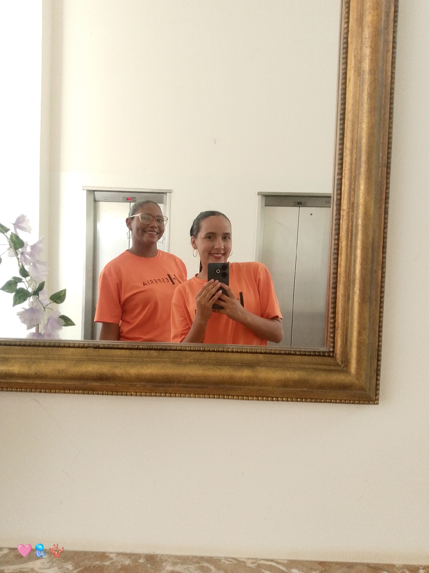 In this full-shot mirror selfie, two young women are captured in a warm and playful moment. The woman on the left, with glasses and a bright smile, wears an orange t-shirt with a reversed logo that reads "AKESCEBA". To her right, the second woman, holding a smartphone to take the picture, also sports an orange t-shirt and a radiant smile. Both women appear to be in good spirits, their joyful expressions suggesting a lighthearted mood.
The setting appears to be an indoor location, possibly a hotel or a public building, hinted by the presence of elevator doors in the background. A large, ornate gold-framed mirror dominates the foreground, reflecting the subjects. To the left of the frame, a branch of delicate purple flowers adds a touch of natural beauty to the scene. The lighting is bright, suggesting it's daytime. At the bottom of the image, three small icons - a pink heart, a blue figure, and a red coral - are visible, adding a touch of whimsy. The overall impression is one of friendship and shared happiness.