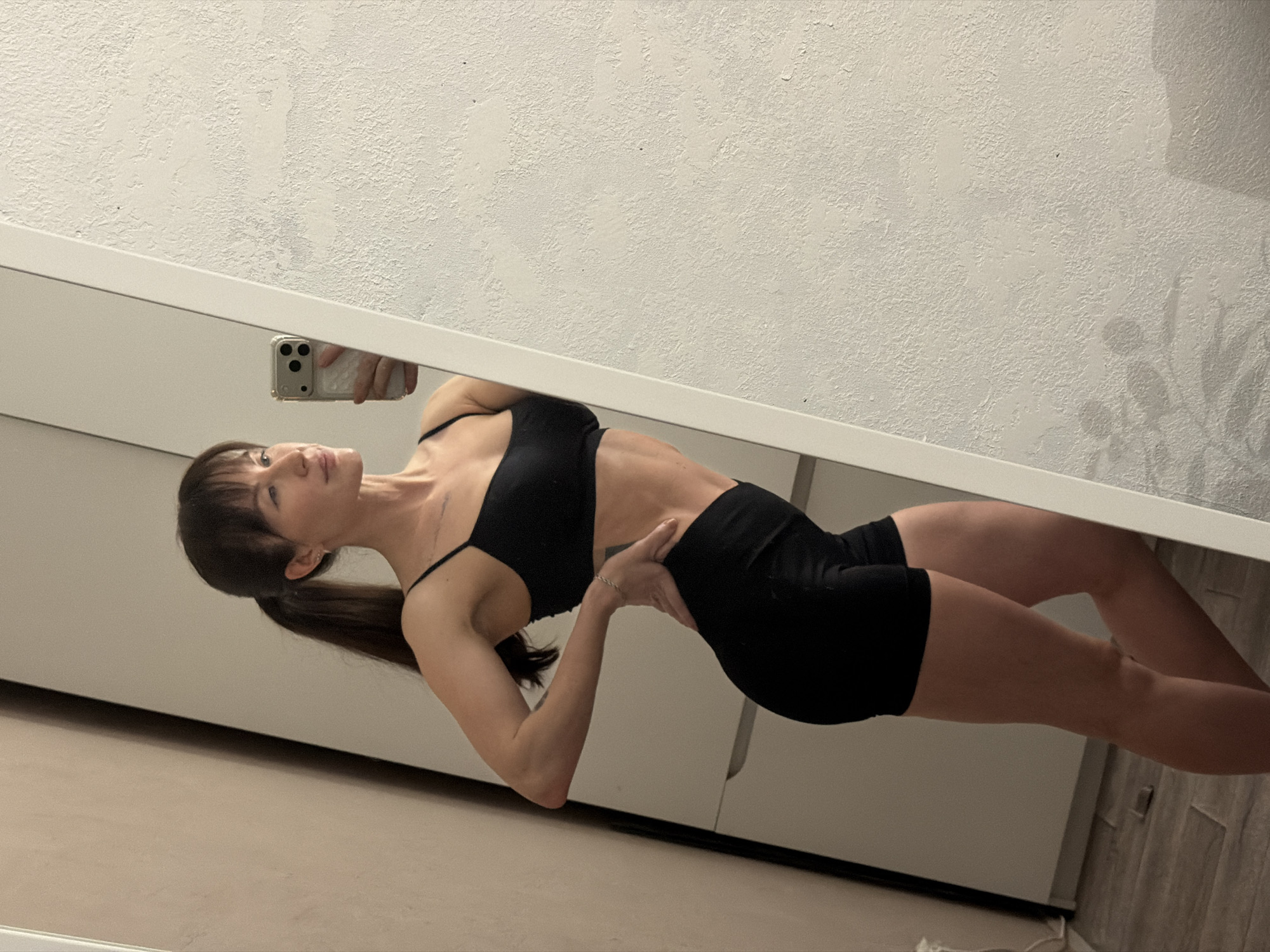A full-body mirror selfie captures a woman posing in an indoor setting, located in Perm, Russia. She has dark hair styled in a low ponytail with bangs and is dressed in a black athletic crop top or sports bra and matching black athletic shorts. Her posture involves a slight arch in her back, with her left hand placed on her lower back, while her gaze is directed upwards and to her right. The reflection shows her right hand holding a light-colored smartphone, equipped with three prominent camera lenses and a textured, possibly quilted, phone case.
The mirror itself features a clean white frame. The environment visible in the reflection above the mirror is a white wall characterized by a distinct, heavily textured finish, possibly stucco or a similar rough plaster, with minor dark flecks. Below the mirror, a light-toned floor and a portion of white, minimalist furniture, likely a cabinet or wardrobe, are discernible. The scene is illuminated by consistent artificial lighting, indicating an indoor environment without strong natural light sources. There is no visible text in the image. The overall impression is a posed picture showcasing fitness or attire within a contemporary home interior.