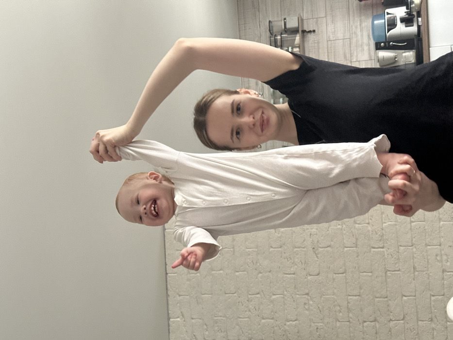 An indoor photograph shows a young adult female holding a toddler aloft. The female, wearing a black short-sleeved top, has her left arm raised, gripping the child's right hand, while her other hand supports the child's bare feet from below. She looks directly at the camera with a slight, neutral expression. The toddler, dressed in a white long-sleeved romper with snap buttons, displays an open-mouthed smile, showing small teeth. Its left hand forms a "thumbs up" gesture, pointing towards the left. The background on the left features a white brick-patterned wall. To the right, a kitchen area is visible, including a wooden shelf with clear food storage containers, a light-colored countertop, and various appliances such as a silver coffee maker and a blue and white blender. The precise location within the city of Chaikovsky, Russia, is not discernible from the image.