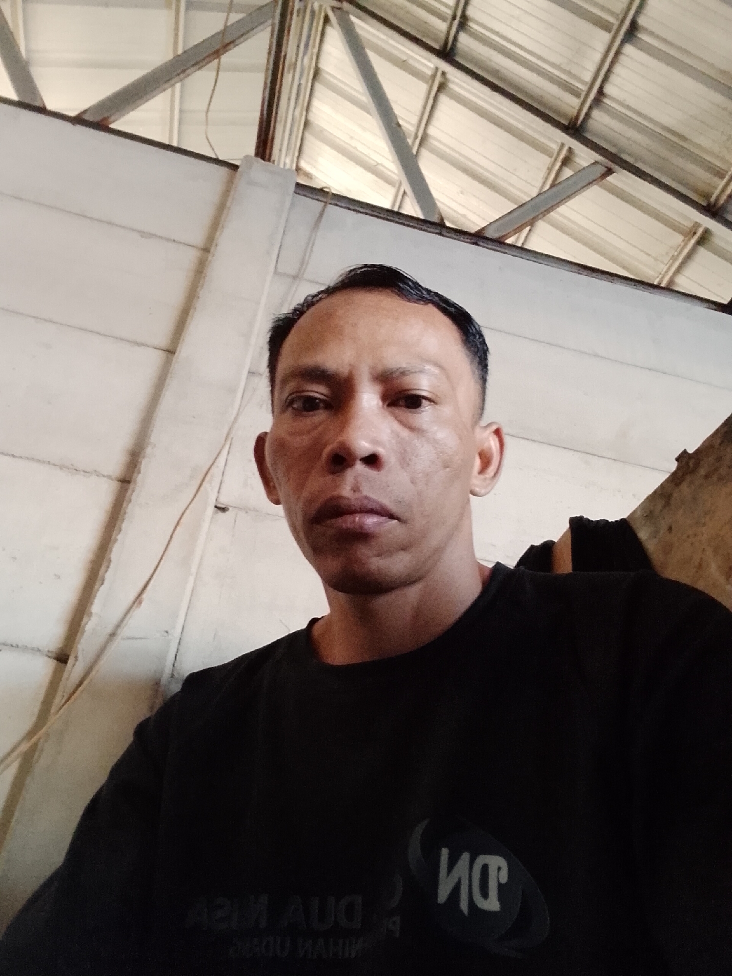A middle-aged man with short, dark hair and a fair complexion is looking at the camera. He is wearing a black t-shirt. The background shows the interior of a building with a corrugated metal roof and exposed beams. There is also a white wall and a section of a brown, textured wall visible. The scene is likely an indoor space, possibly a workshop or storage area, in Bukit Ketok, Indonesia.