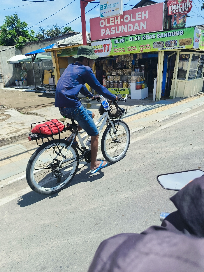 The image shows a man in a blue shirt and blue jeans riding a white bicycle on a paved road in Padalarang, Indonesia. The man is wearing a blue cap and flip-flops. He is carrying a red bag on the back of the bicycle. There are buildings on both sides of the road, one of which is a store with a sign that reads "Oleh-Oleh Pasundan" and "Oleh-Oleh Khas Bandung." The location based on coordinates is in a neighborhood with shops and businesses, a typical urban setting. The man is moving away from the camera on the right side of the road. The road is lined with a white curb. In the foreground, a black motorcycle with a rear-view mirror is facing the camera, and the lower half of its rider, obscured by the motorcycle, is wearing a black jacket. The background is a clear blue sky. The scene is captured under bright sunlight, creating clear shadows.