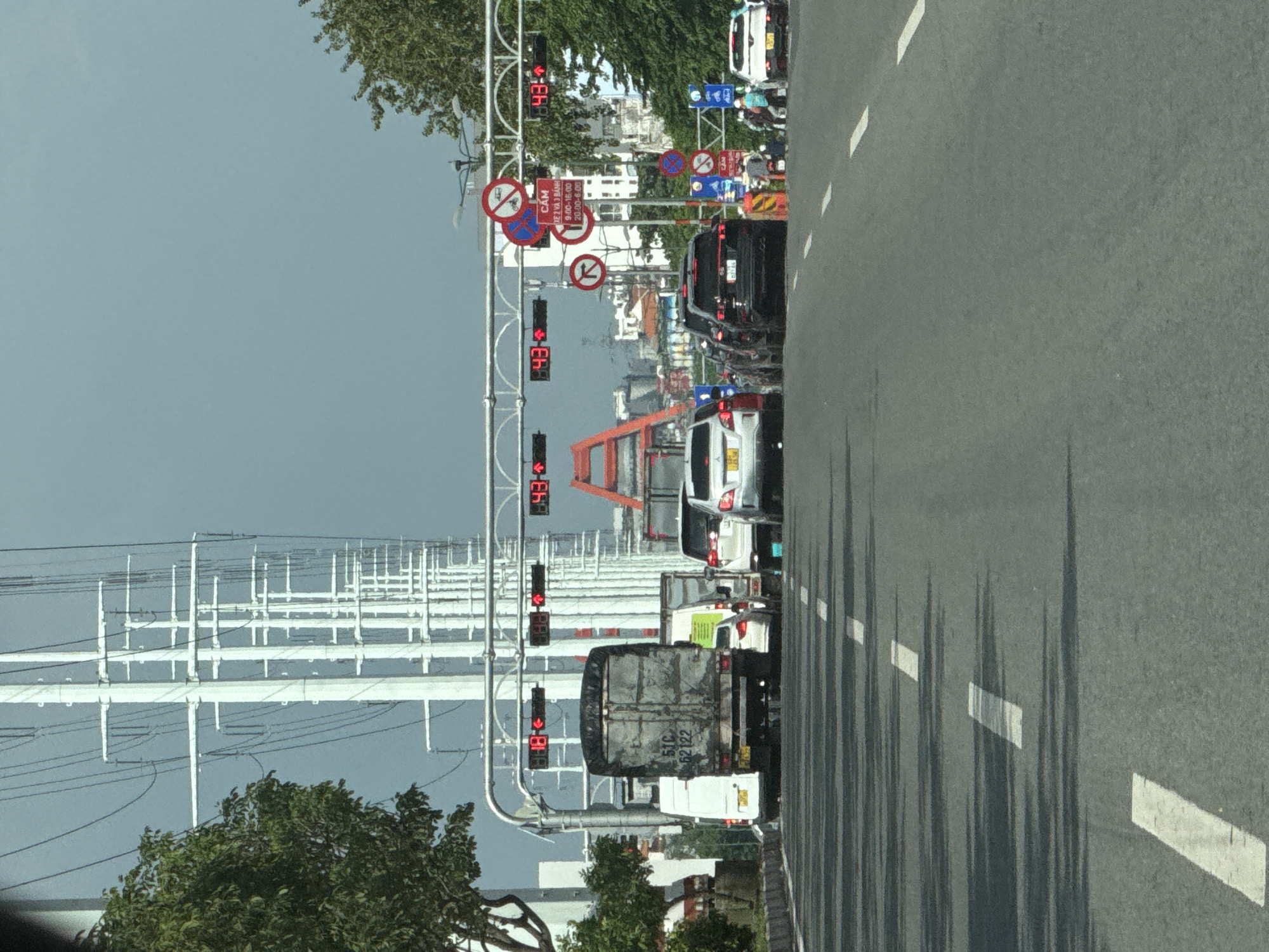 In Thủ Đức City, Vietnam, a traffic jam is captured on a multi-lane road under a grey sky. A line of vehicles, including trucks, vans, and cars, are stationary on the asphalt. Overhead, a complex network of power lines stretches across the scene, supported by tall white poles. Traffic lights displaying red arrows and numerical countdowns are visible above the roadway, indicating waiting periods. Several traffic signs are also mounted on poles, some featuring red circles with diagonal bars, prohibiting certain actions, and others with Vietnamese text, likely related to traffic regulations. Trees with green foliage line the roadside, and in the distance, buildings and parts of a bridge structure are discernible. A few pedestrians and motorcyclists are present on the side of the road, observing the traffic situation.