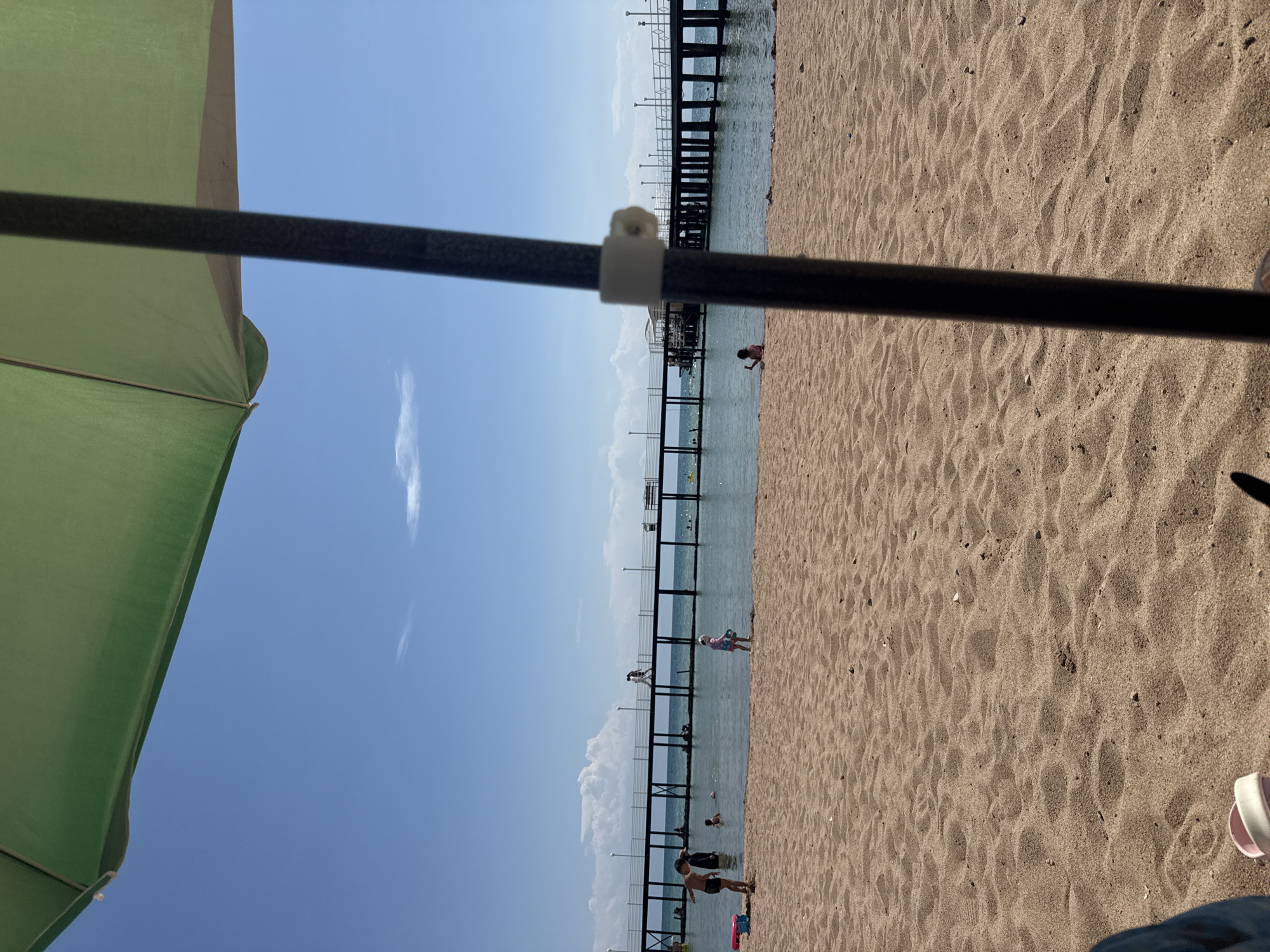 An outdoor scene on a sunny day at a beach in Sary-Oy, Kyrgyzstan. A large green umbrella occupies the top left portion of the frame, casting a shadow over the foreground. The beach is covered in light brown sand, with small shells scattered across it. In the middle ground, several people are visible in the shallow water and on the sandy shore. A long pier stretches across the water in the background, lined with lampposts and a railing. The sky is clear blue with a few wispy clouds.