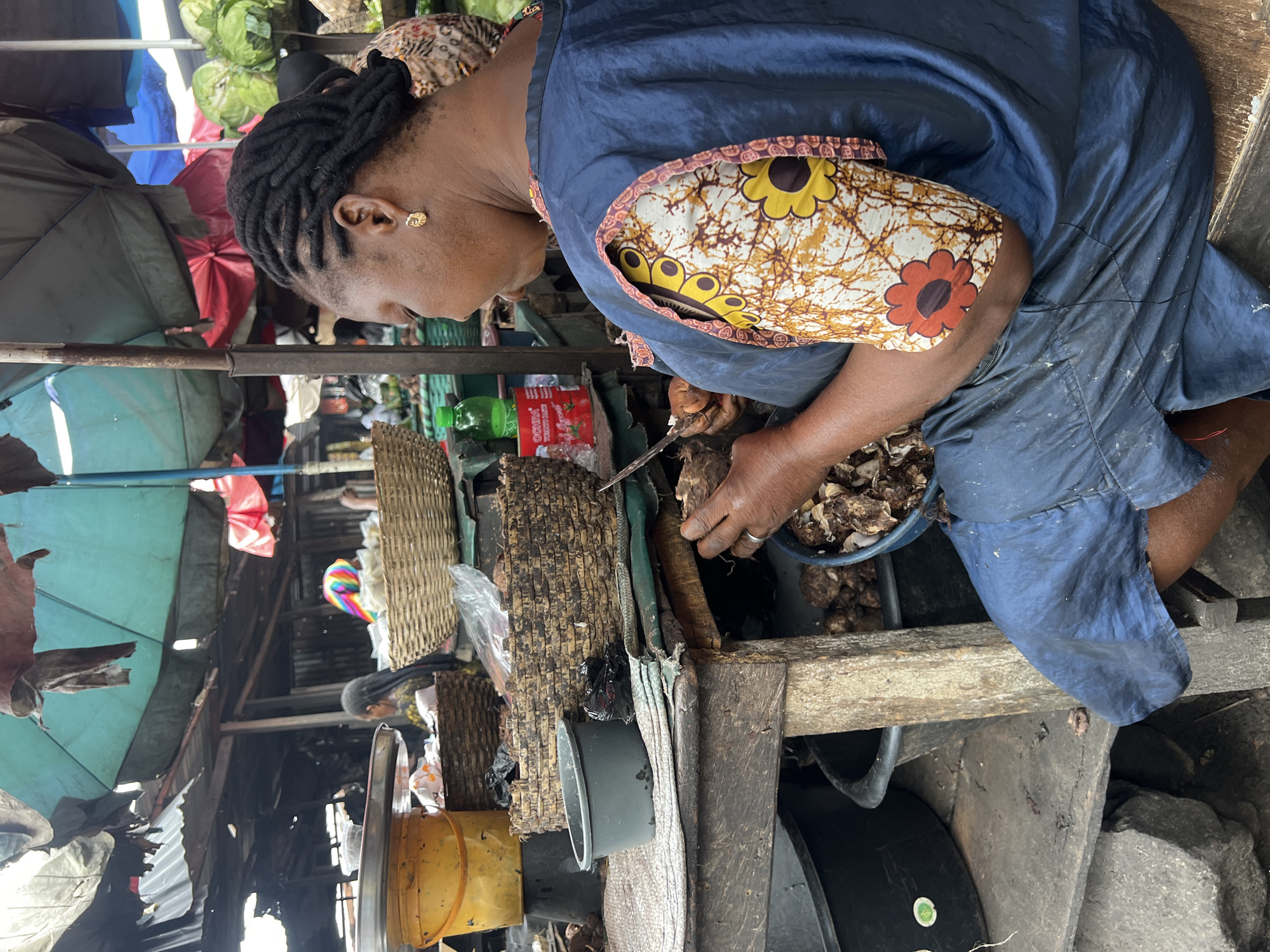 An adult female in Nkpor, Nigeria is seated at a makeshift wooden table in an outdoor marketplace. She is wearing a blue dress with a patterned orange and yellow fabric inset. Her hair is styled in dark braids. She is peeling a root vegetable using a knife. A blue plastic basin nearby holds peeled pieces of the vegetable. Behind her is a cluttered scene of market stalls, including baskets and covered merchandise. A red OCHHA tomato paste packet is partially visible. The overall setting is a somewhat impoverished urban market environment.