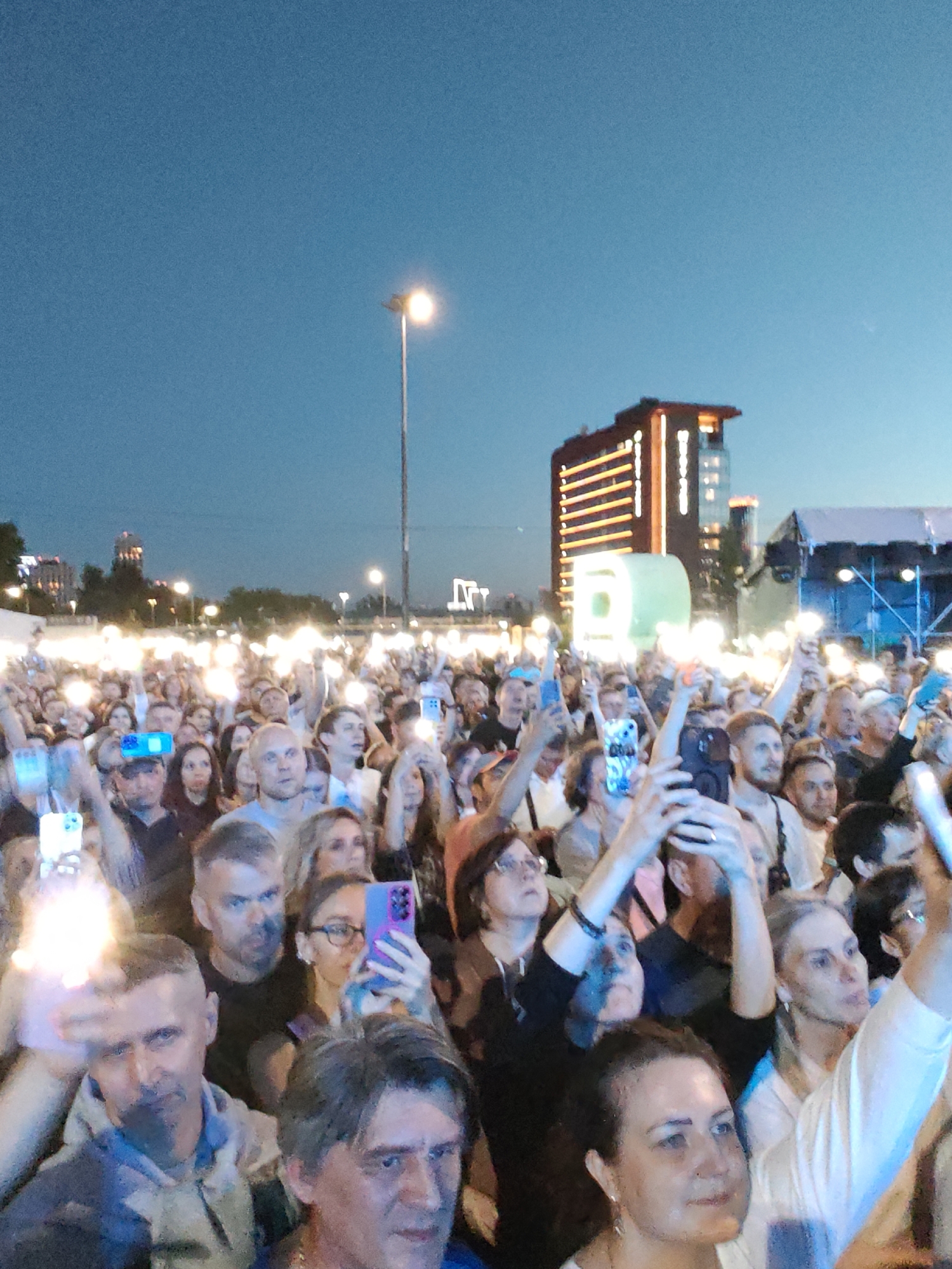 A large crowd of people are gathered outdoors at dusk, with many holding up smartphones with illuminated screens. In the background, a modern, multi-story building with illuminated windows is visible, as well as stage lighting and tents. The scene appears to be a concert or festival. The presence of the building and stage infrastructure, combined with the outdoor gathering and dusk lighting, suggests a public event likely taking place in a city. Given the context of Yekaterinburg, Russia, this scene could be depicting an event at a venue such as the Ekaterinburg Central Stadium or a similar outdoor entertainment space.