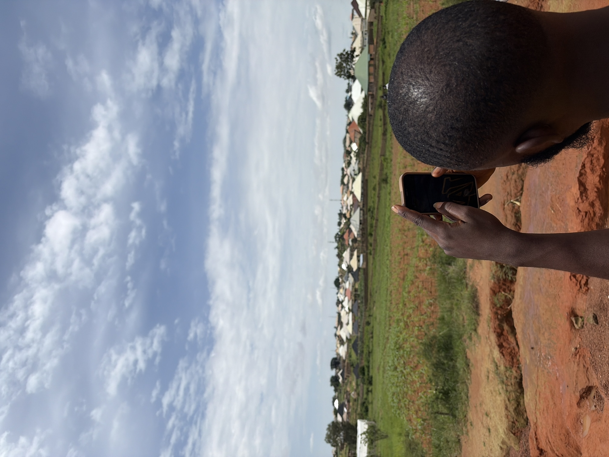 In Rafin Bauna, Nigeria, a person is captured from behind, holding a smartphone in their left hand and pointing it towards a village settlement. The individual's head and the back of their neck are visible, with short, dark hair and a beard. They are positioned on a reddish-brown embankment, with a field of green crops and scattered red soil patches in the mid-ground. The background is dominated by a sprawling village with numerous buildings featuring white and light-colored roofs, some with darker, conical roofs. A few trees are scattered throughout the settlement and the surrounding landscape. The sky above is partly cloudy, with formations of white clouds against a blue backdrop. The overall scene depicts a rural or semi-rural setting with a focus on the village and the person's interaction with it through their phone.