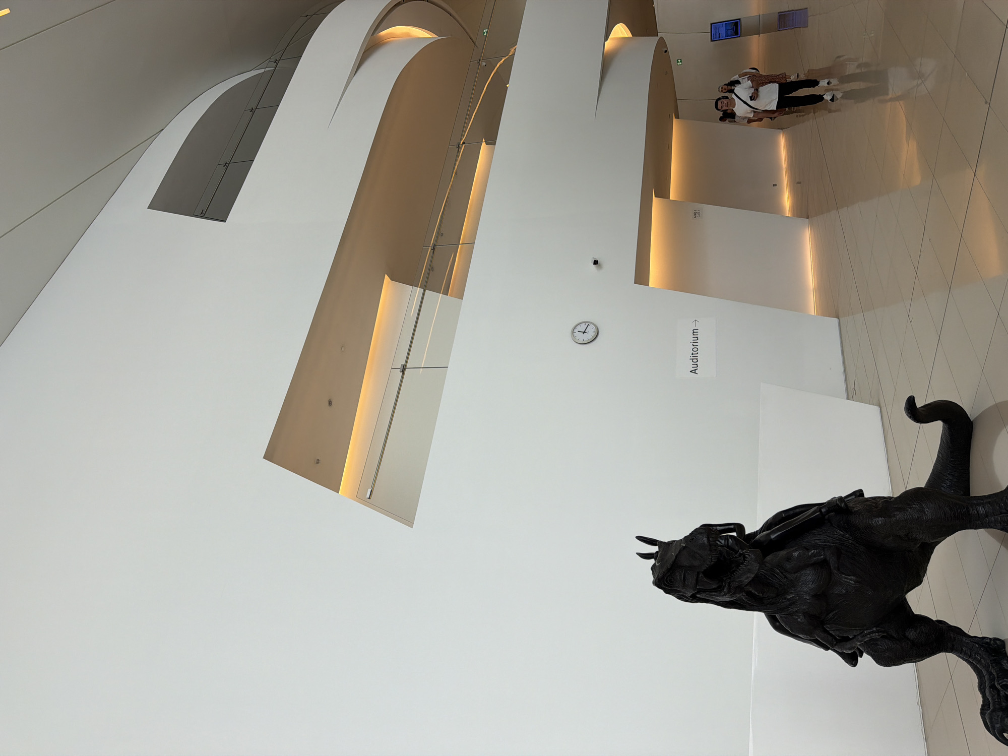 The image depicts the interior of a modern building, likely a museum or gallery, in Baku, Azerbaijan. The architecture is characterized by smooth, white, curved walls and ceilings with subtle, warm lighting accentuating the architectural lines. In the foreground, a dark, detailed sculpture of a dinosaur-like creature with a figure riding on its back stands on a polished floor. To the right of the sculpture, a sign points towards an "Auditorium." Further into the background, several people are walking through the brightly lit hallway. One man in a white t-shirt and black pants, carrying a black bag, walks towards the camera, while other individuals are visible in the distance. The overall impression is one of contemporary design and public space.
