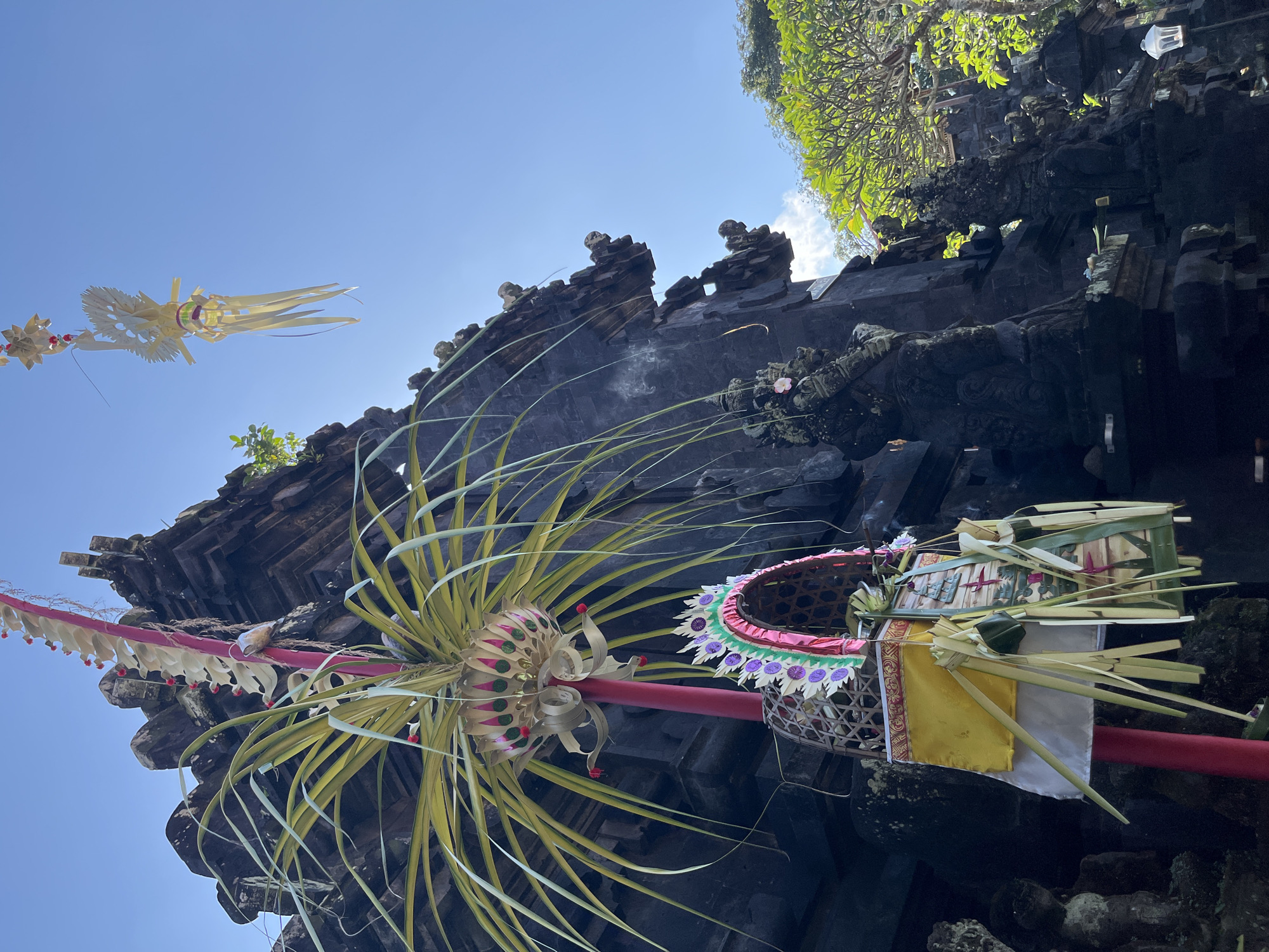 The photograph depicts the entrance to a dark stone temple in Sukawati, Indonesia, on a clear day. Intricate Balinese decorations, including palm frond creations and offerings, adorn a red pole in the foreground. The temple structure features weathered stone carvings of mythical figures. Lush green trees and a bright blue sky are visible in the background.
