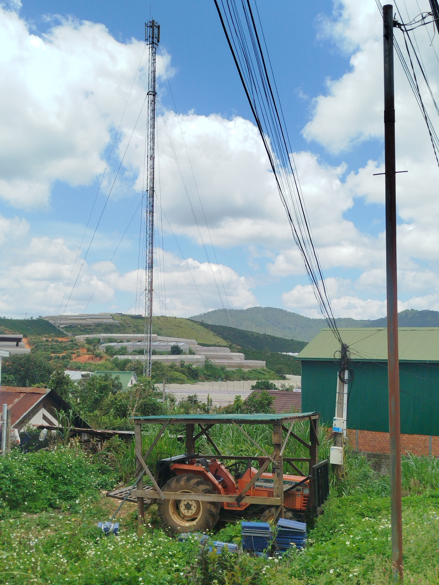A rustic scene unfolds in Thôn 6, Vietnam, under a bright, cloudy sky. In the foreground, a red tractor sits under a makeshift wooden shelter with a green tarp roof. It is surrounded by lush green grass and white wildflowers. To the left of the shelter, a small house with a rusted metal roof is visible, and further back, several other buildings with green roofs are scattered amongst trees. In the middle ground, a tall communications tower stands prominently against the sky, its metallic structure crisscrossed with guy wires. Beyond the tower, rolling hills are covered with numerous greenhouses, suggesting agricultural activity in the area. Power lines run diagonally across the image, supported by a tall, rusty pole on the right side of the frame, with electrical boxes attached to a utility pole near the green building. The overall atmosphere is one of rural life, with a blend of modern infrastructure and natural landscape.