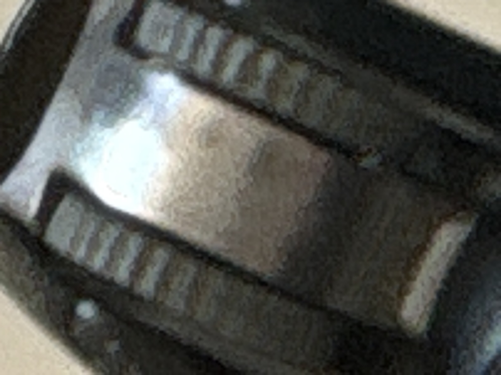 The image is a close-up, abstract view of a dark, metallic object with textured surfaces. Two ribbed or grooved sections are visible on either side of a central, smooth, and reflective surface that appears to be a lighter color, possibly silver or a brushed metal. The lighting suggests a strong light source reflecting off the central area, creating highlights and shadows. The overall impression is one of manufactured precision and a slightly industrial aesthetic. There are no discernible elements of a cityscape or environment.