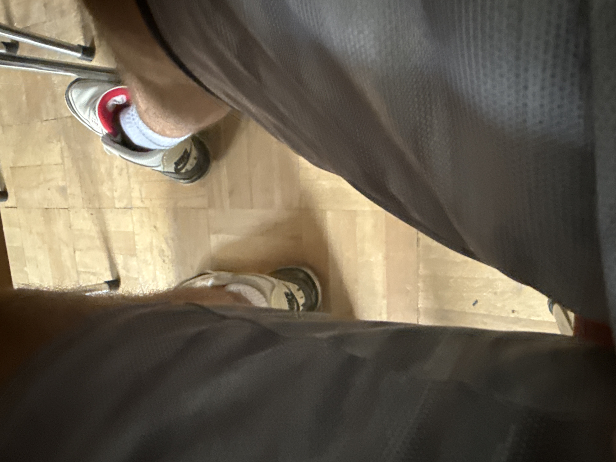 The image shows a close-up view of two people's legs and feet, sitting on what appears to be a wooden floor. Both individuals are wearing athletic shoes and shorts. One person is wearing white socks with red accents in their shoe, while the other's socks are not clearly visible. The lighting suggests an indoor setting, possibly a home or a casual public space. The image does not provide any specific visual cues that would allow for its placement in Sofia, Bulgaria.