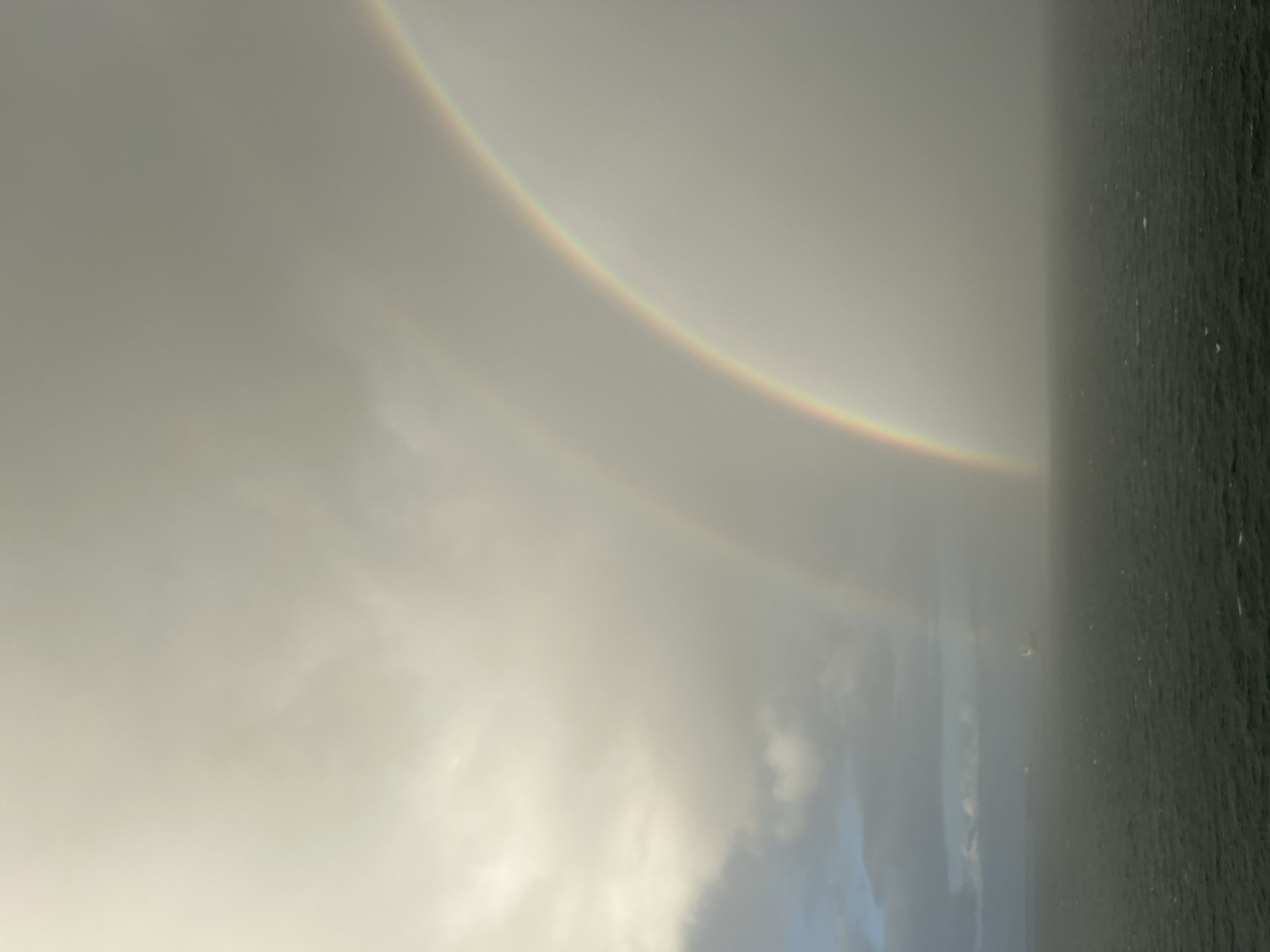 The image depicts a hazy scene with a double rainbow visible in the sky. The foreground is dominated by mist or low clouds obscuring the ground. To the right, there is a body of water. The location appears to be near Niagara Falls, Unknown.