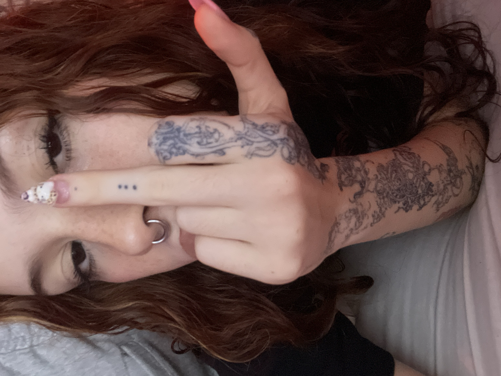 A close-up, tilted portrait features a person with long, wavy reddish-brown hair lying down, likely on a bed or soft surface, indicated by light-colored bedding and a pillow. The subject's left eye, adorned with dark eyelashes, is partially visible, along with a portion of their nose, which has a silver hoop septum piercing. Freckles are noticeable on their face.
Their right hand is raised in the foreground, partially obscuring the lower half of their face. The middle finger is extended, and the thumb is raised, forming an 'L' shape. The thumb nail is long and painted light pink, while the middle finger nail features a decorative pattern. Extensive, intricate dark-colored tattoos with ornate designs cover the back of the hand and extend up the forearm. Three small, dark dots are tattooed on the side of the extended middle finger.
The person is wearing a dark garment. The lighting is soft and ambient, typical of an indoor setting. The scene conveys a casual atmosphere. No discernible text is visible in the image. The provided location context is Buenos Aires, Argentina.