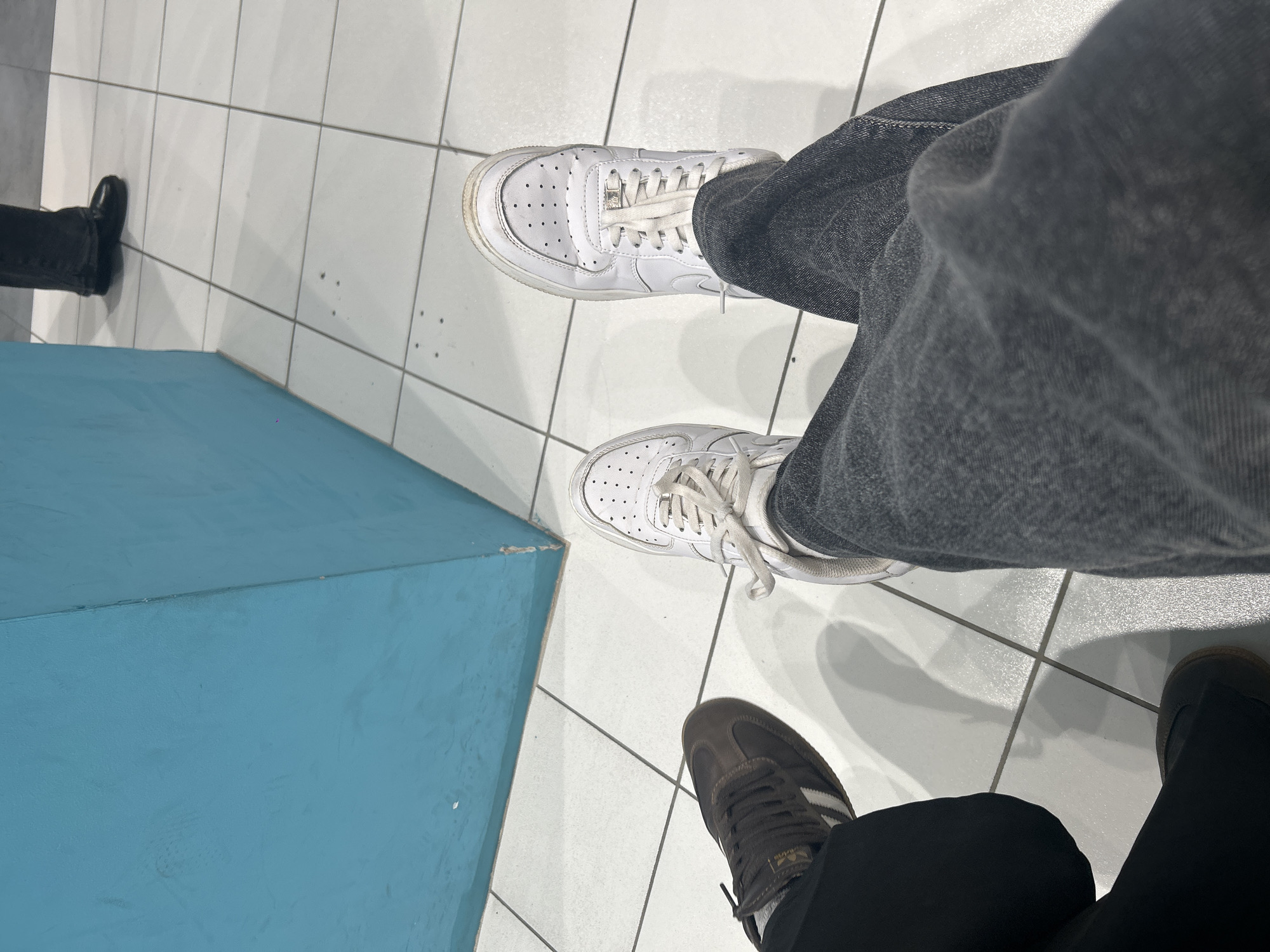 This top-down, slightly rotated image captures the feet of at least two individuals standing on a light-colored tiled floor with dark grout lines.
The primary subject wears light-washed or grey denim jeans and white Nike Air Force 1 sneakers. These sneakers show visible signs of wear and dirt, particularly around the edges and toe box. A small, metallic lace lock is present on the laces.
Adjacent to this, another person's foot is visible, wearing dark pants and dark brown Adidas Samba sneakers, distinguished by their iconic three white stripes on the side and a light-colored tag on the tongue. Shadows from the people's legs and shoes are cast on the tiled floor, indicating an overhead light source.
In the upper-left corner, a third person's foot, clad in a black formal-style shoe and dark pants, is partially visible next to a large, solid blue, rectangular block or structure. This blue object occupies a significant portion of the left side of the frame and shows minor surface imperfections.
The setting appears to be an indoor public space, such as a mall or waiting area, located in Saint Petersburg, Russia. The people are standing, and no specific activity or event is clearly depicted beyond this. No legible text is visible on the footwear or other objects.