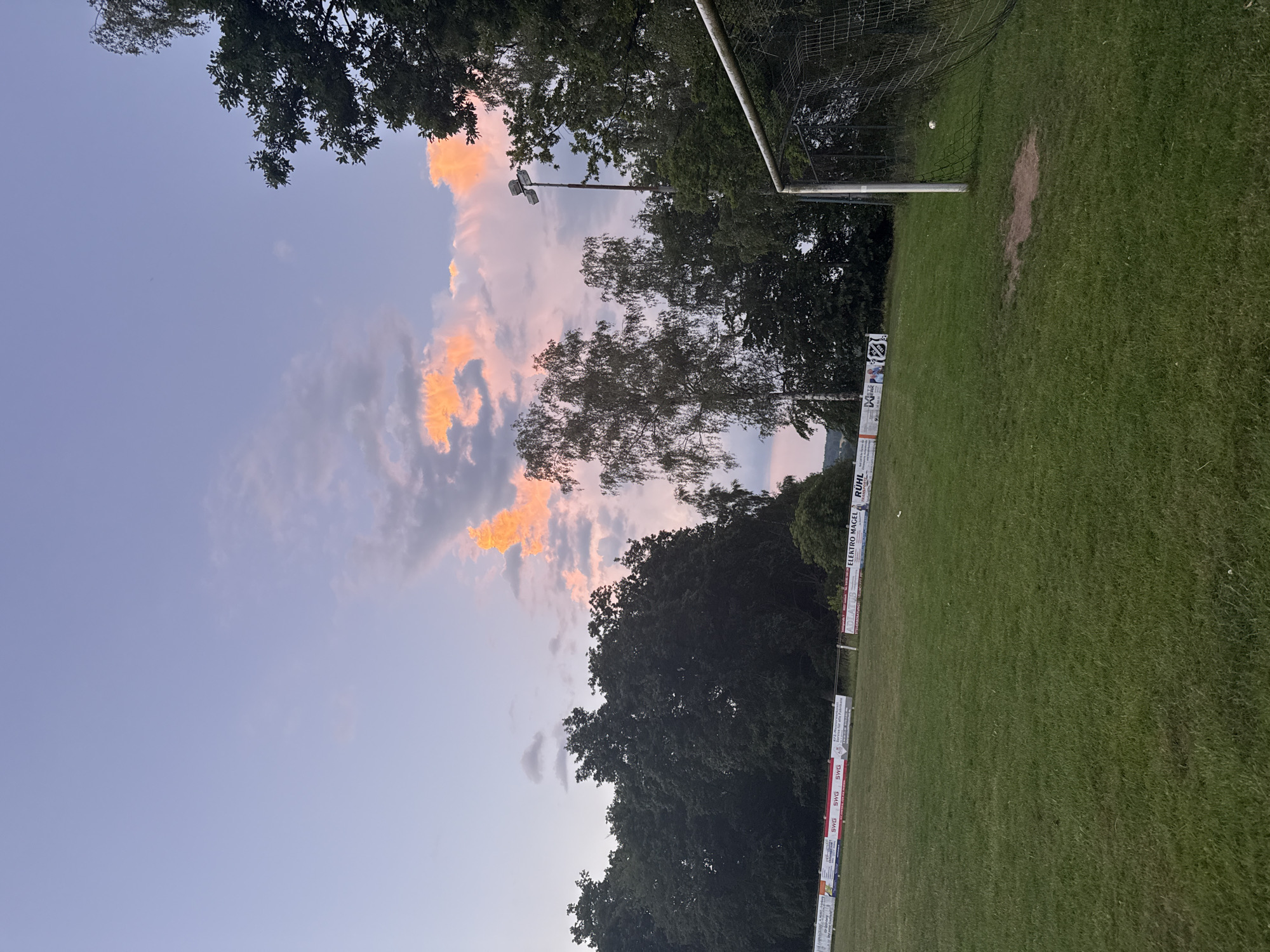 The image depicts a soccer field in Lumda, Germany, during twilight. The sky is a gradient of soft purples and blues, with scattered clouds catching the last light of the sun, tinged with orange and pink. In the foreground, a well-maintained green grass field stretches towards a soccer goal on the right. The goal is a standard metal frame with netting, and a white soccer ball rests inside the net. Along the far side of the field, a line of trees forms a dark, dense backdrop. A row of advertising banners is visible behind the trees, some featuring German text and logos, including "SWG". A tall lamppost with multiple lights stands near the trees and the goal. The overall atmosphere is serene and quiet, suggesting the end of a day or a break in activity.